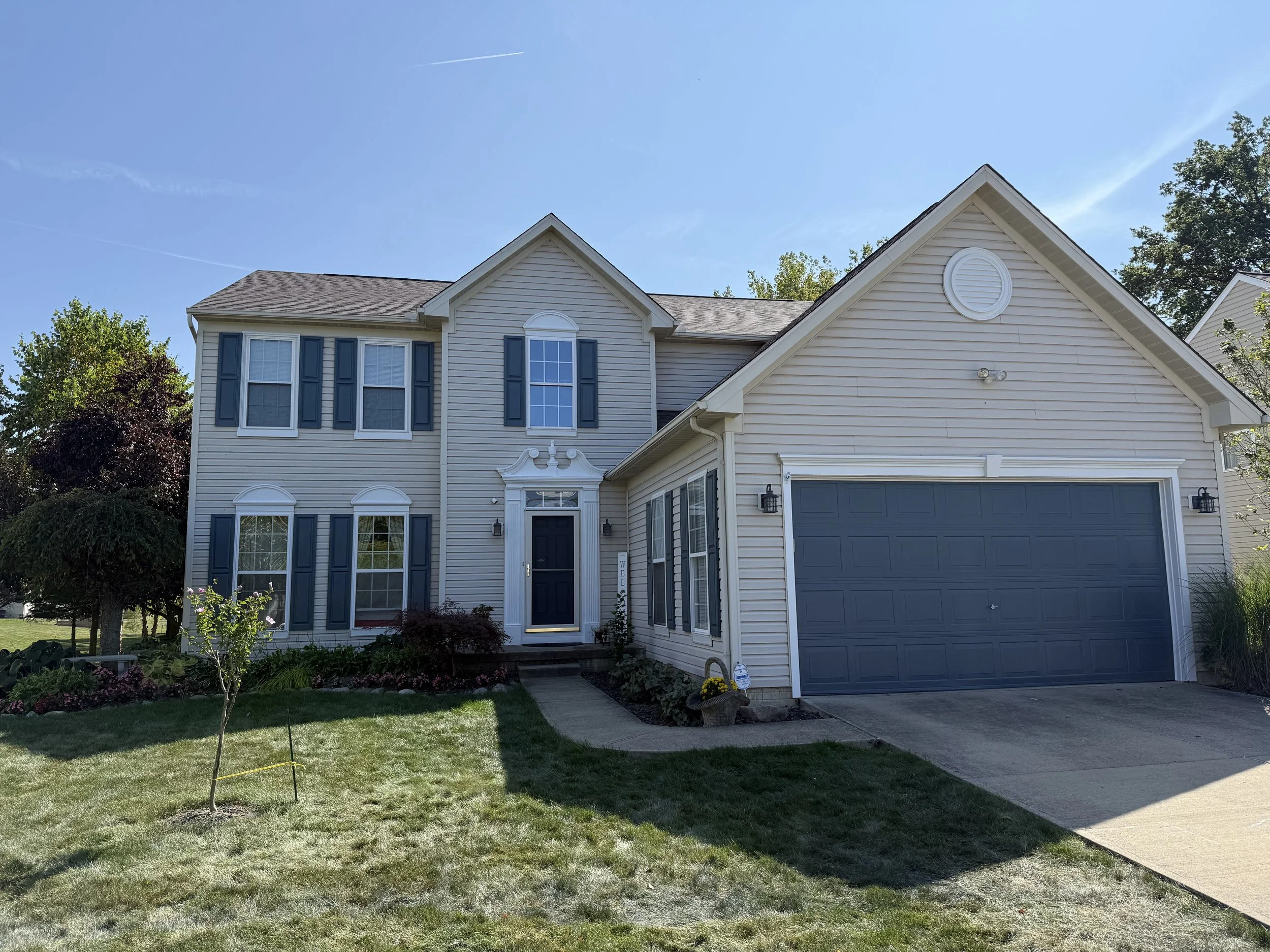 A two-story suburban house with beige siding, dark blue shutters, and a dark blue garage door. The house has multiple white-trimmed windows, a front door with a decorative pediment, and a small front yard with grass, shrubs, and a young tree. The driveway leads to the garage, and there are exterior lights on either side of the garage door.