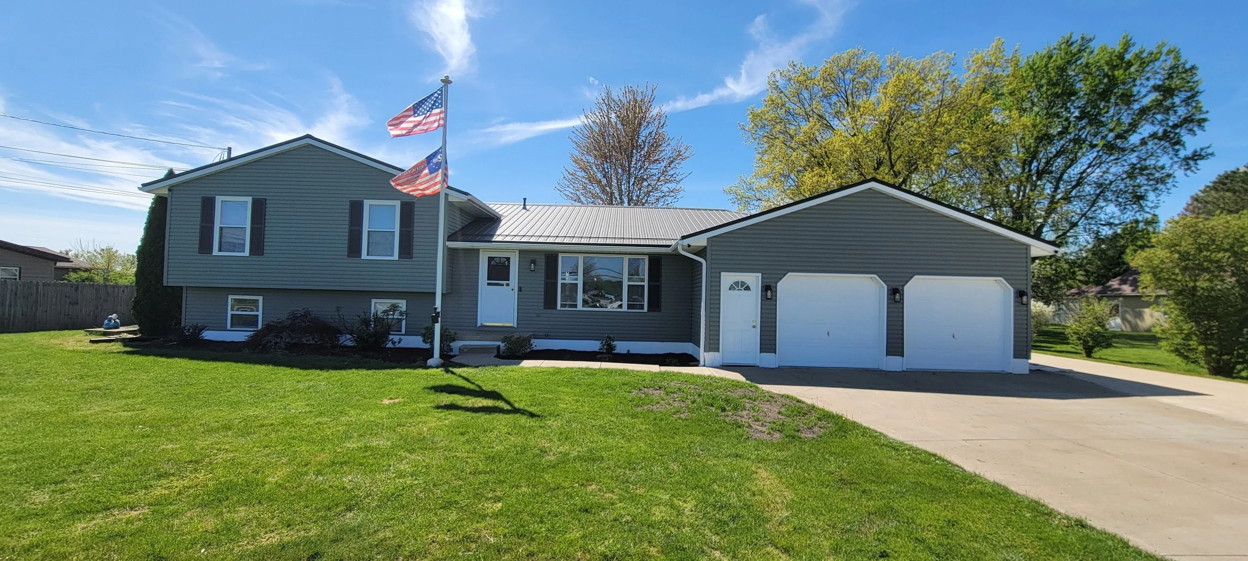 A two-story gray house with a attached two-car garage, American flags on a flagpole, a green lawn, and trees in the background, under a blue sky.