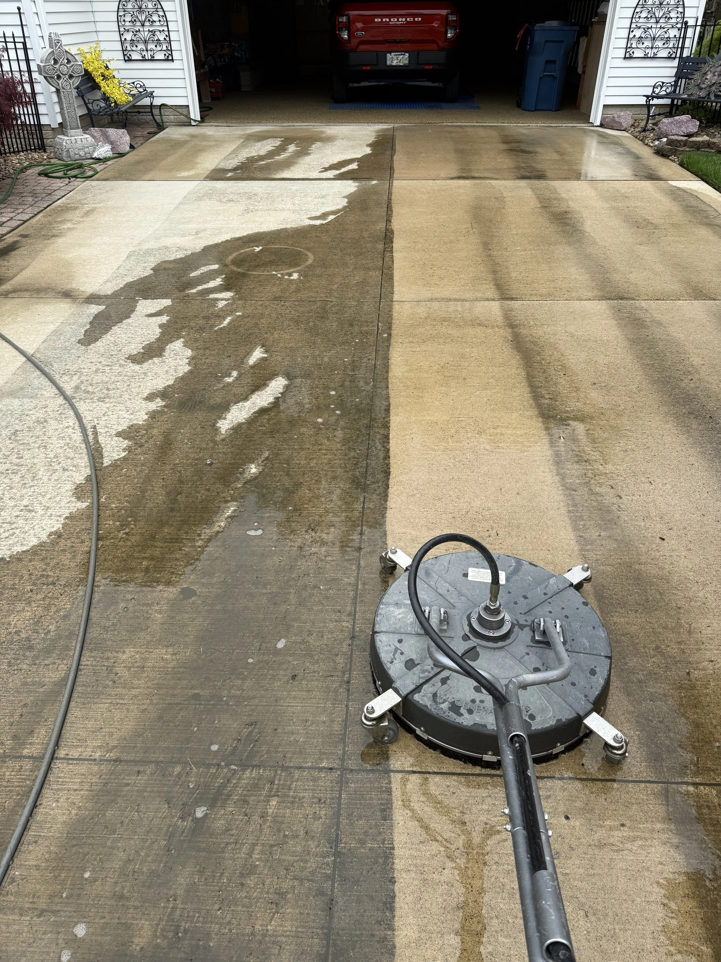 A pressure washing machine is cleaning a concrete driveway, with water and soap visible on the surface. The driveway leads to a garage with a red truck parked inside, and houses with decorative elements are visible on the sides.
