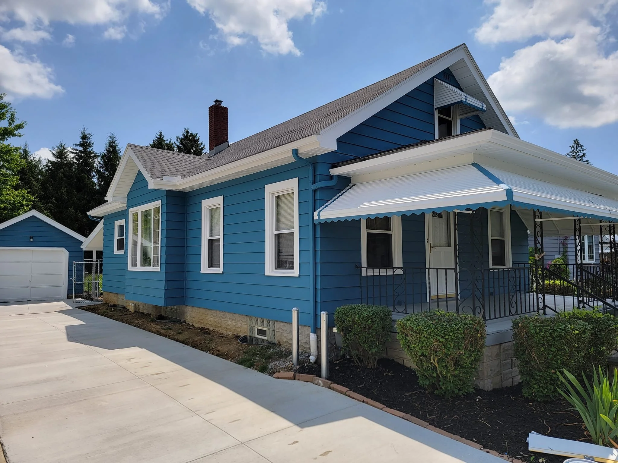 Blue house with white trim, front porch with decorative iron railings, front yard with shrubs, concrete driveway, detached garage in the background, partly cloudy sky, surrounded by green trees.