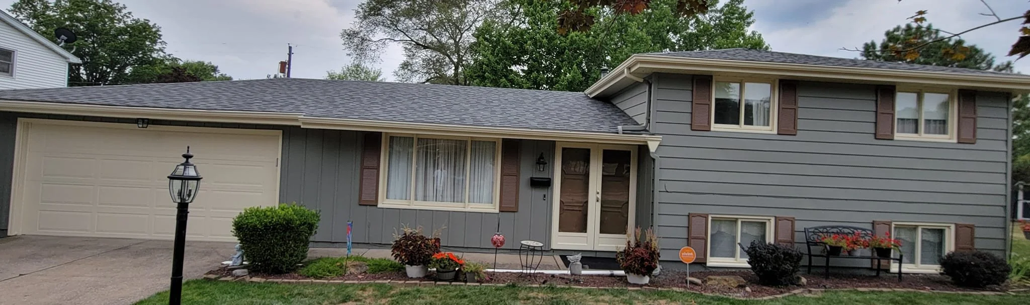 A gray two-story house with a front yard, small garden, and outdoor bench, featuring beige window shutters, a lamp post, and a brick-lined flower bed.