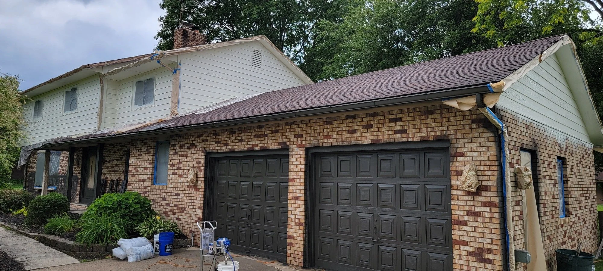 A two-story brick house with a black garage door, a partially covered driveway, and window protectors indicating ongoing renovation.