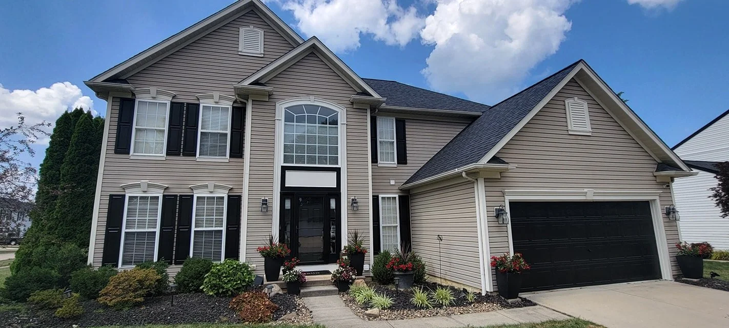 Front view of a two-story house with beige siding, black shutters, and a black garage door, surrounded by landscaped garden beds with shrubs and flowers.