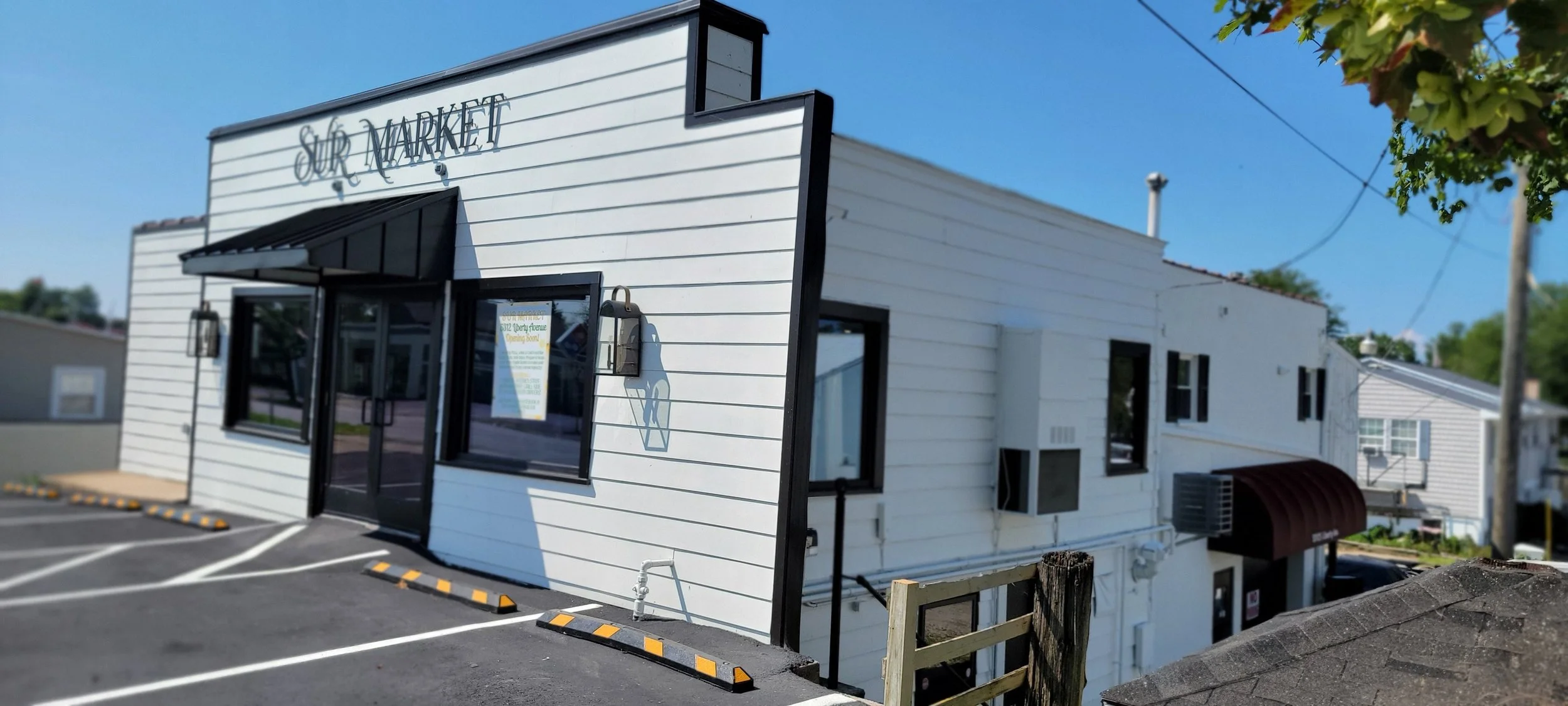 White building with black trim and a sign that reads 'SUR MARKET' in cursive, with a parking lot in front and a blue sky above.
