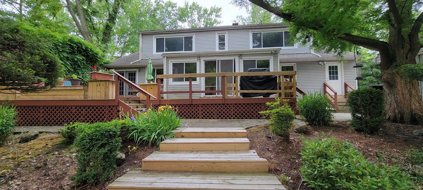 Backyard view of a two-story house with a wooden deck, stairs leading to a garden, and lush greenery including bushes and trees.