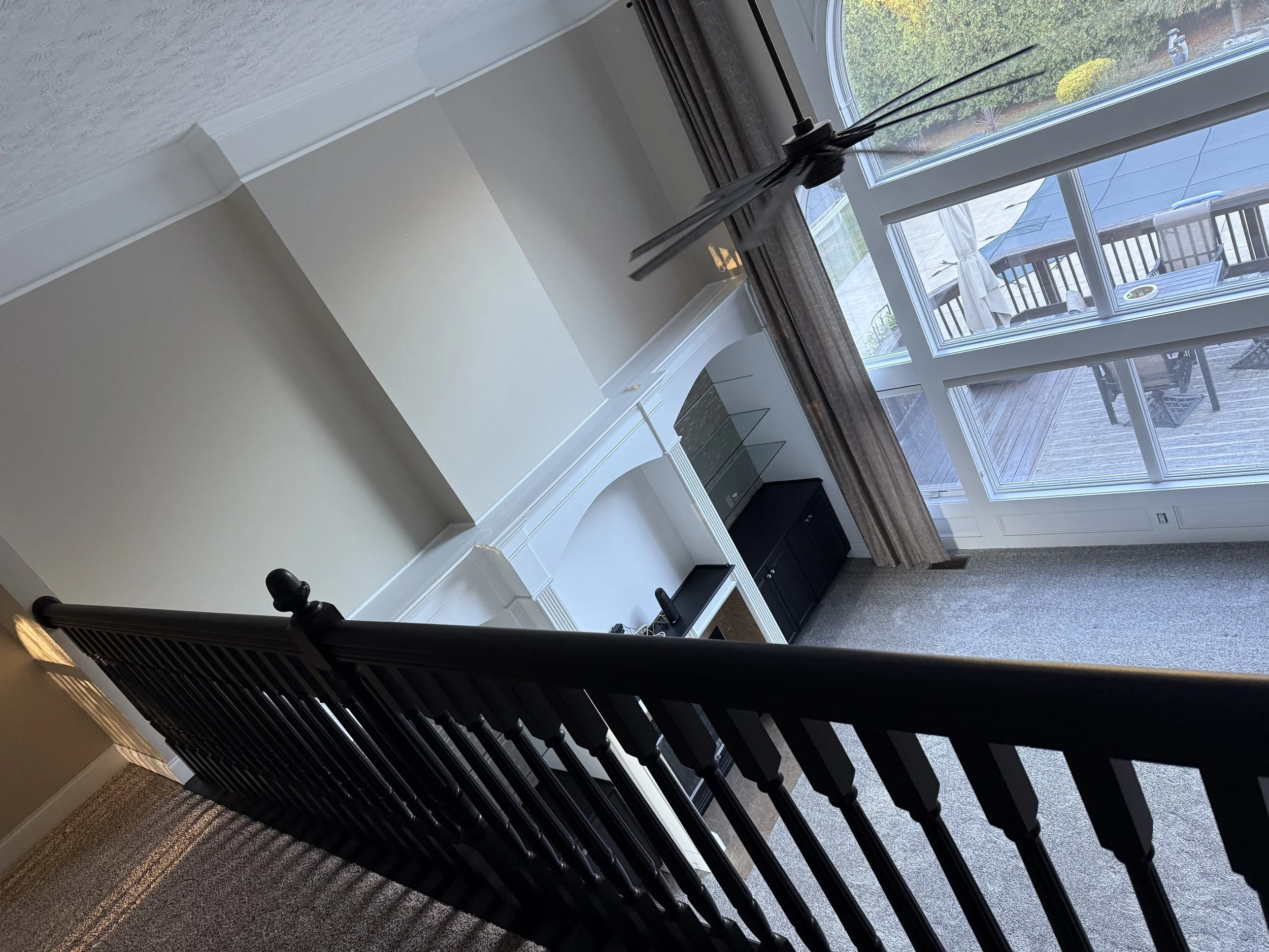 A staircase with a black wooden banister leading to a room with beige carpet. The room has a large window and glass door revealing an outdoor patio with a table, chairs, and umbrellas. There is a white fireplace mantel with glass shelves and black ca
