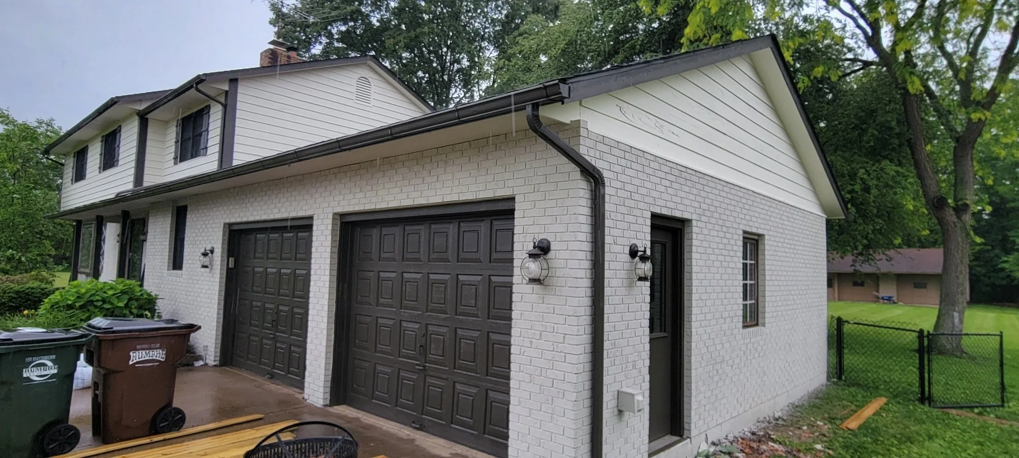 A two-story house with white brick exterior and black garage doors, surrounded by green trees and lawn, with outdoor lighting fixtures beside the garage and a black mesh fence on the right.