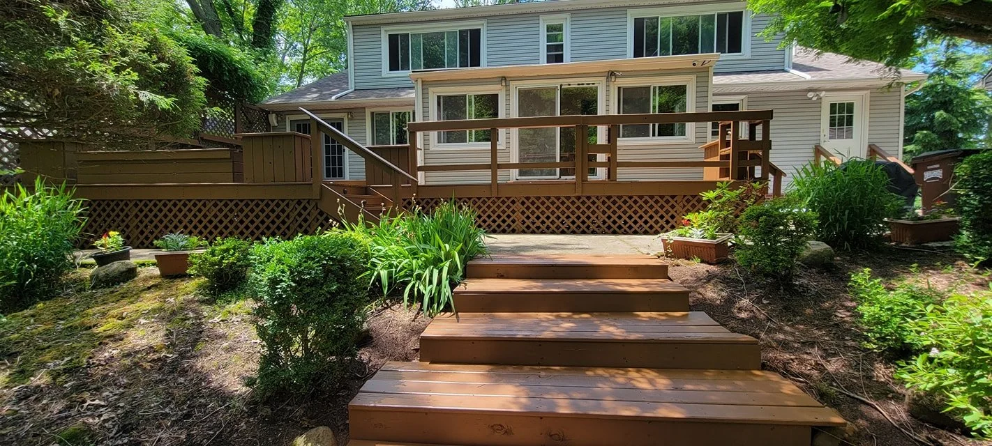 Backyard with a wooden deck, stairs, and a house in the background, surrounded by trees and greenery.