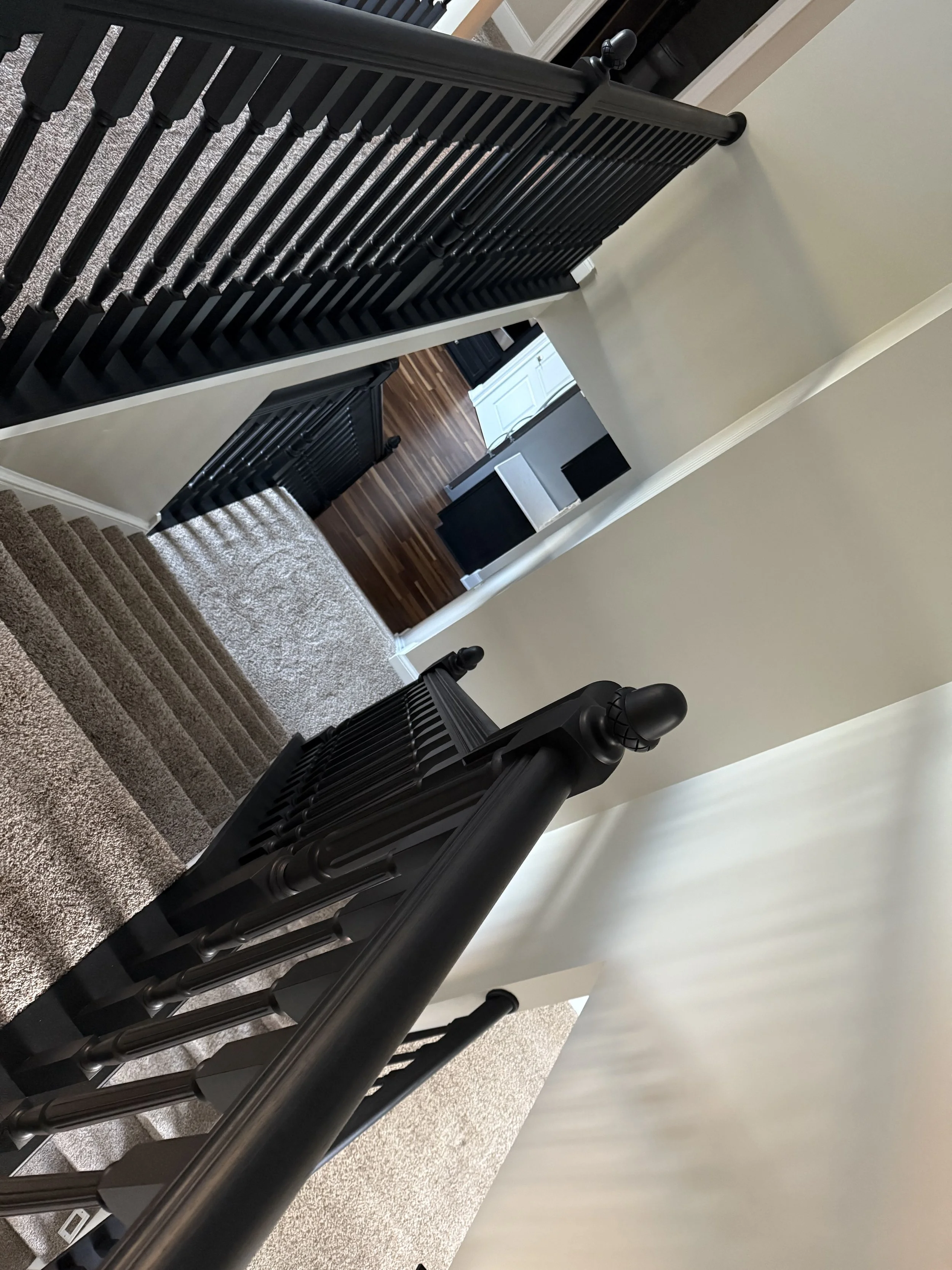 View of a staircase with black balusters and handrails, carpeted stairs, beige walls, and a view into a room with dark wood flooring and furniture.