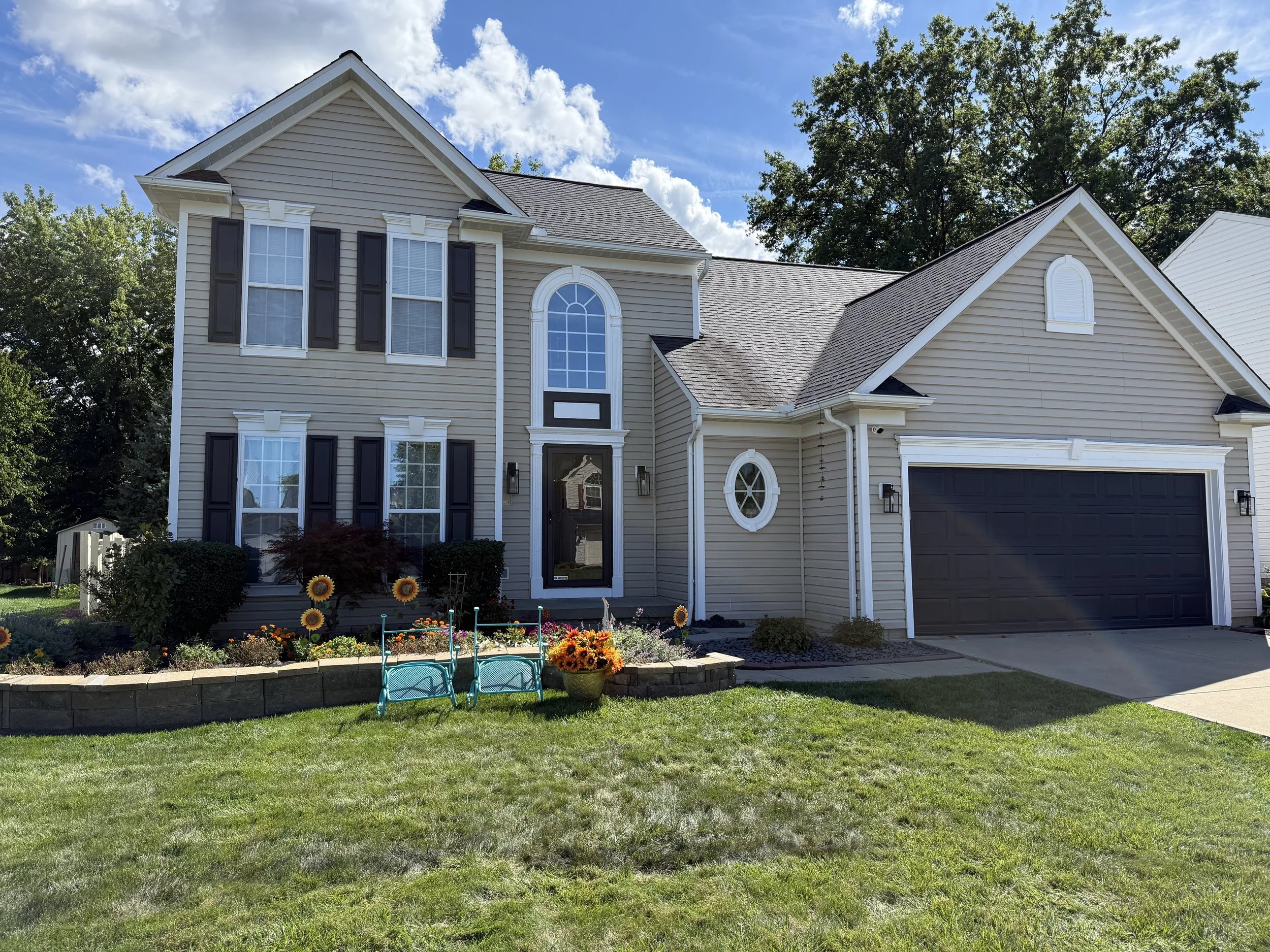 Front view of a two-story house with beige siding, black shutters, and a black garage door, surrounded by a green lawn and a flower bed with sunflowers and other plants, under a partly cloudy sky.