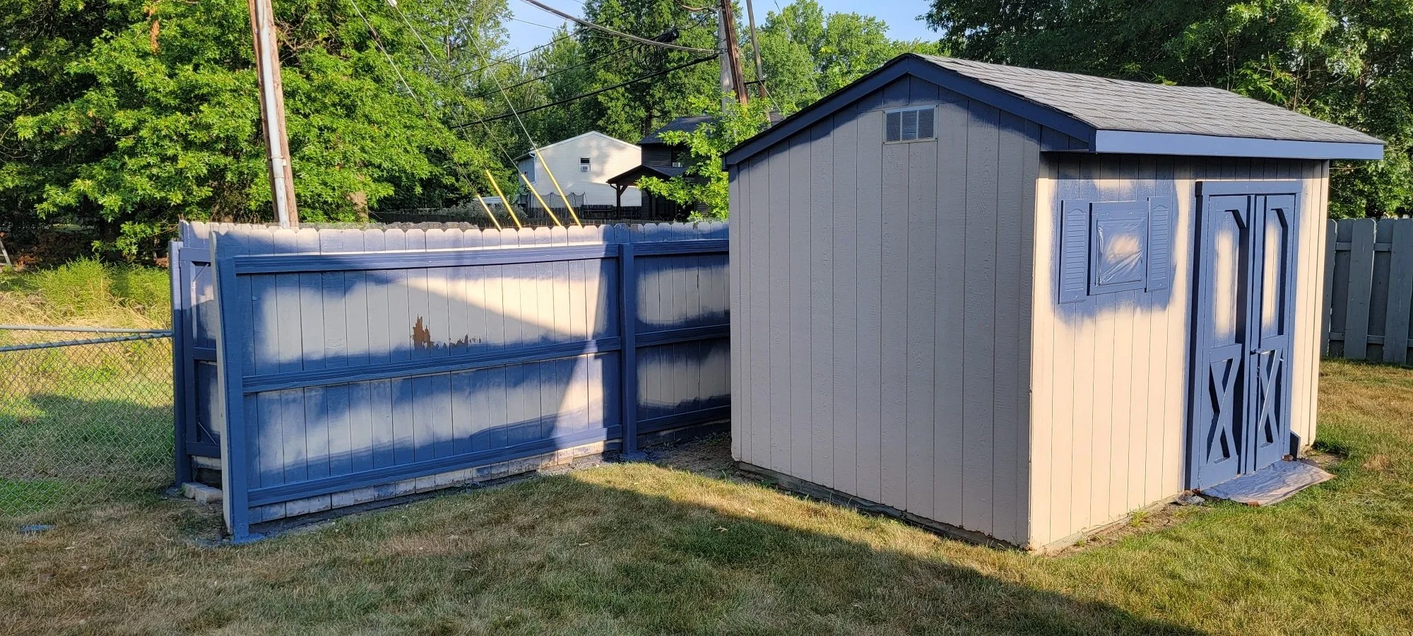 A backyard scene with a small light gray storage shed with blue double doors, a window with a shutter, and a blue fence with a chain-link section. There are trees and houses in the background, and the grass is green.