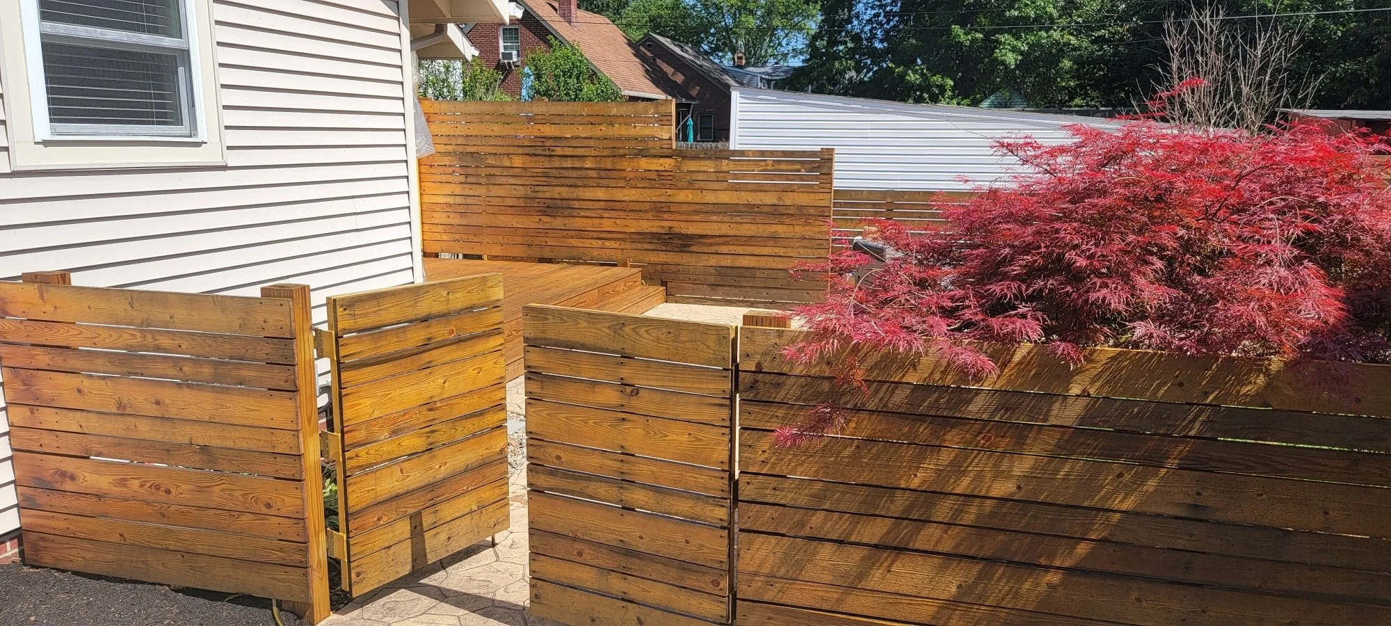 A backyard with wooden fences, a white house wall, a window, and a red flowering tree.