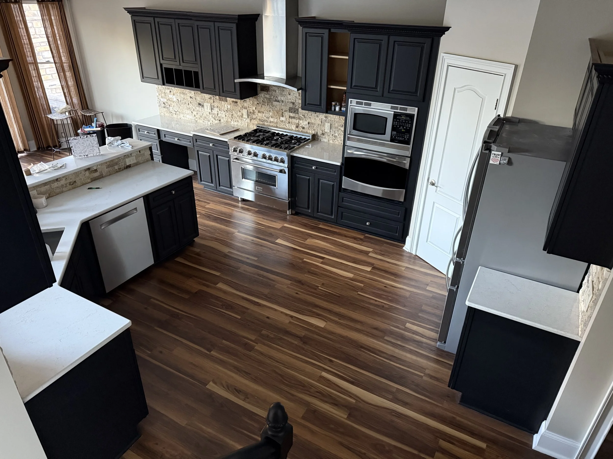 Modern kitchen with black cabinets, stainless steel appliances, a gas stove, microwave, and refrigerator, hardwood floors, and a brick backsplash.
