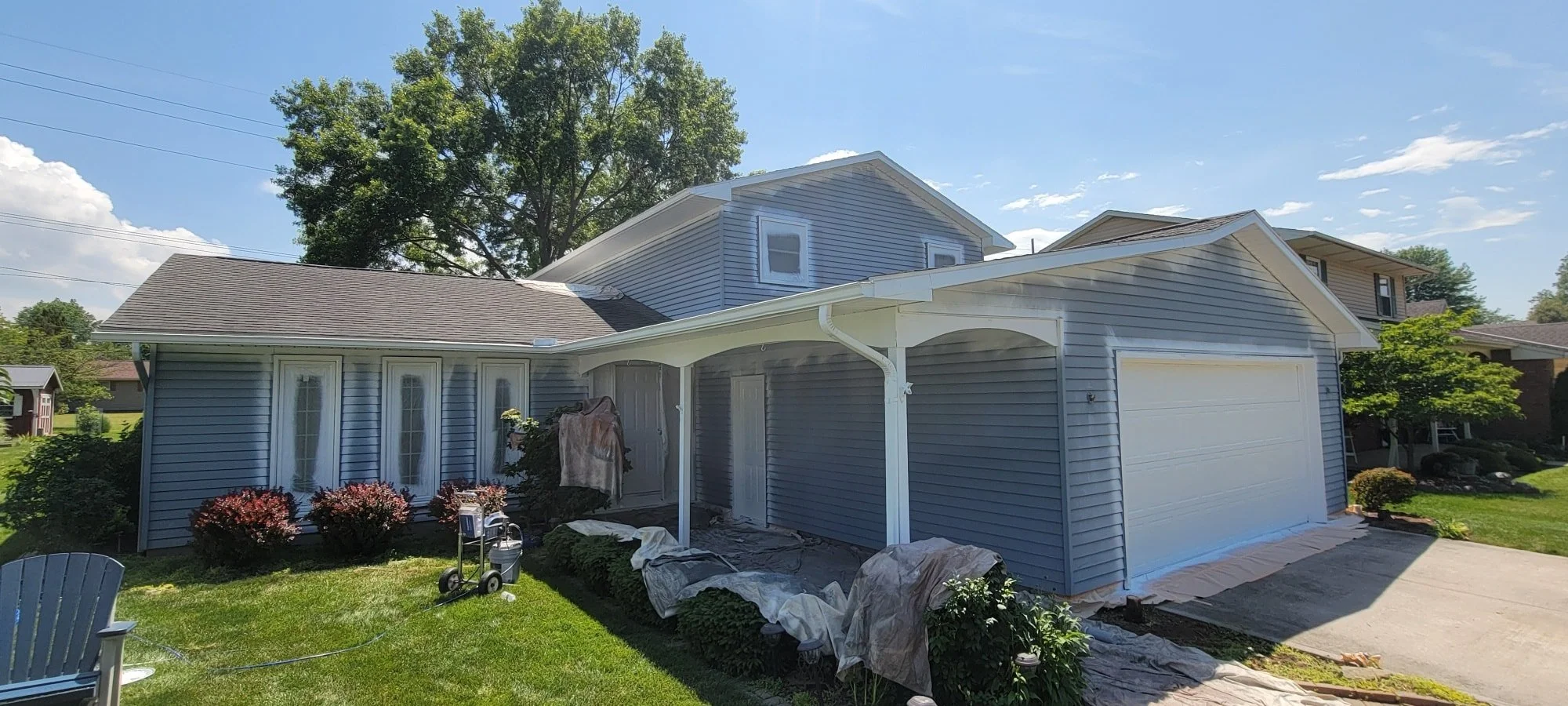 A light blue house with a white garage door and a small covered porch, surrounded by green grass, shrubs, and trees, under a clear blue sky.