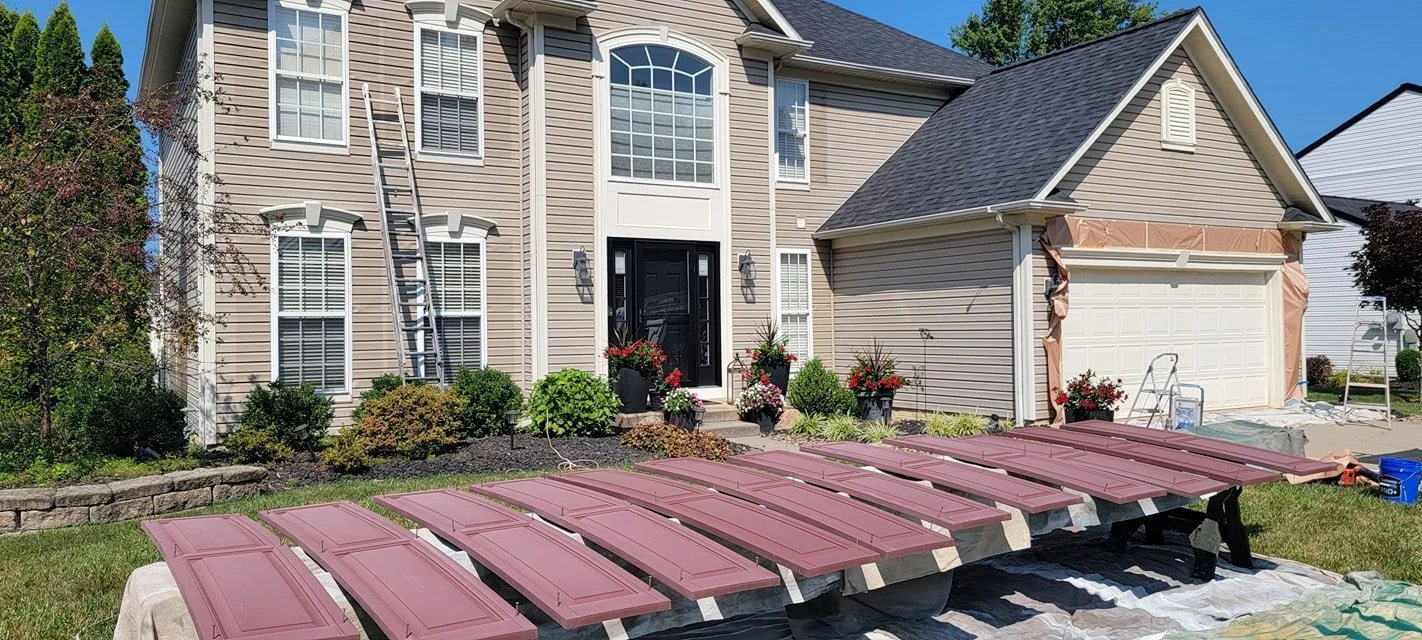 Exterior view of a beige two-story house with a black front door, multiple windows, and a side garage door under construction. Multiple freshly painted maroon cabinet doors are laid out on saw horses in the front yard, with various tools and supplies