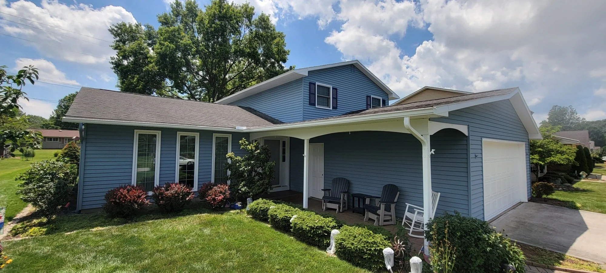 Front view of a two-story house with blue siding, white trim, a white garage door, and a small porch with outdoor chairs. Green lawn with shrubs and flowers, large trees in the background, partly cloudy sky.
