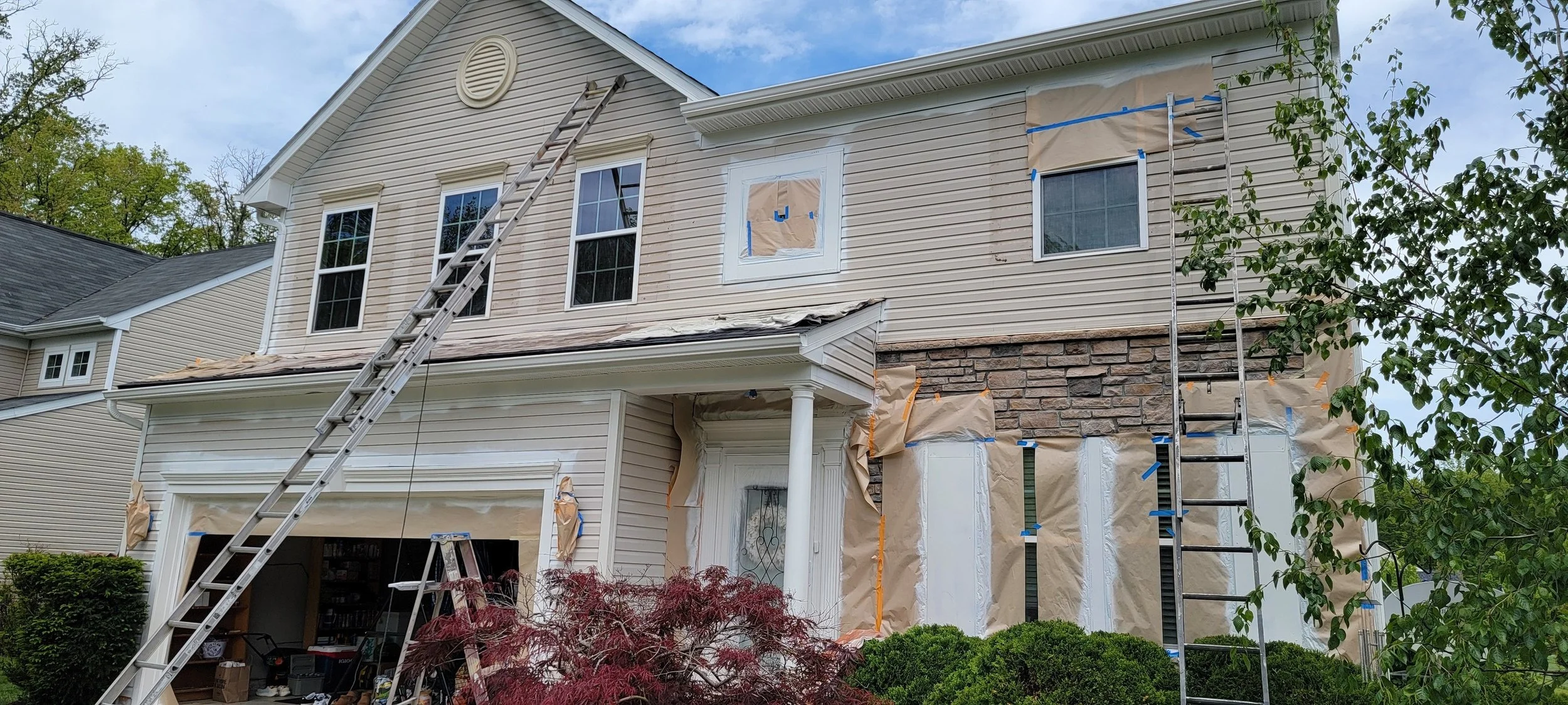 A two-story house undergoing exterior renovation with beige siding and stone accents. Ladders are propped against the house, with windows covered in paper and blue painter's tape. The roof has some damaged or missing shingles, and construction materi