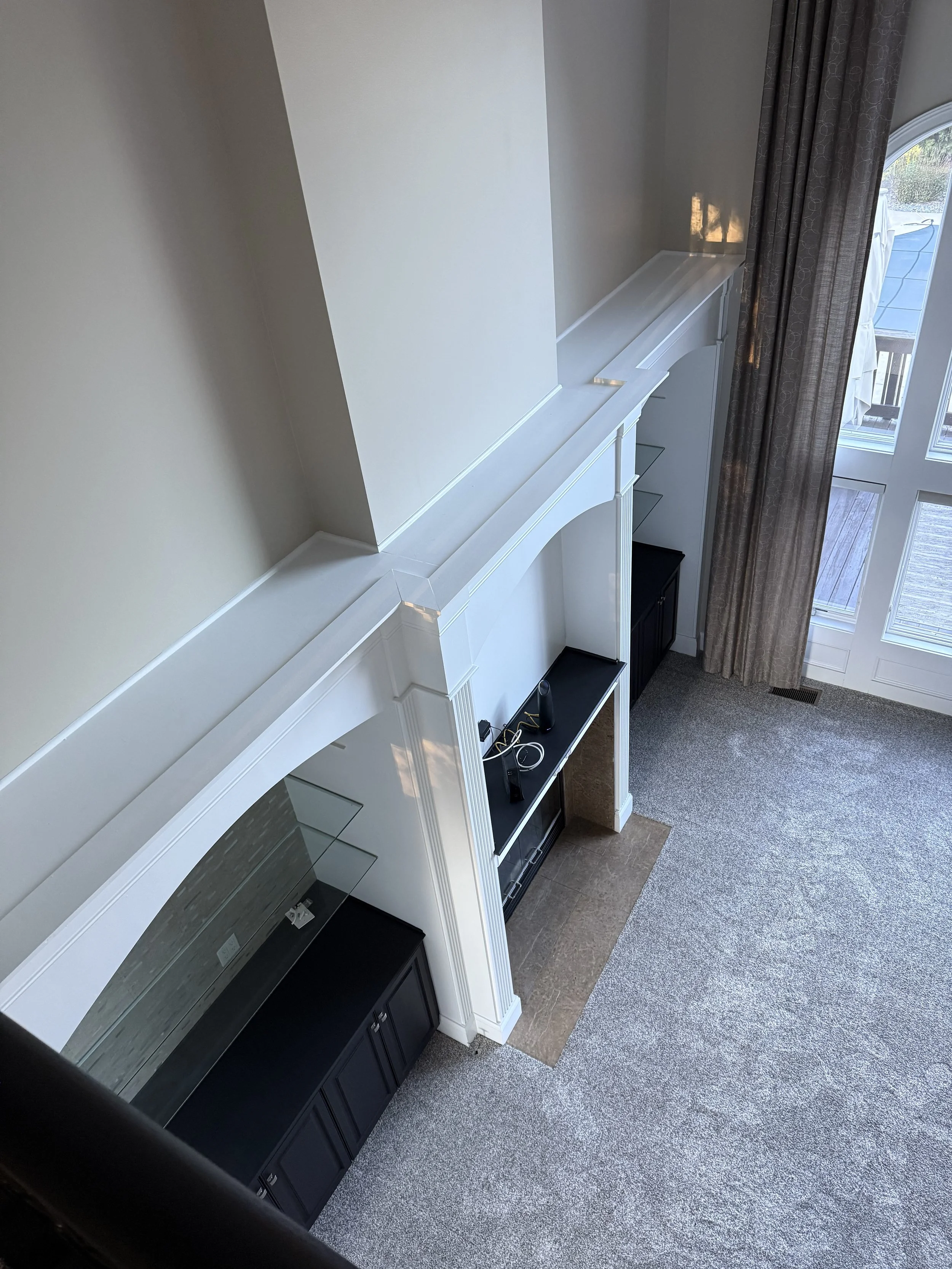 Overhead view of a living room with a white fireplace mantel, black cabinets underneath, and gray carpet, next to a glass door with curtains.