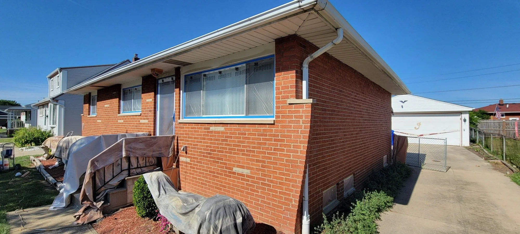 Side view of a brick house with a white roof, several windows covered with protective plastic, and a driveway leading to a garage with a diamond-patterned door. There's gardening and outdoor furniture covered with cloth outside the house. Clear blue 