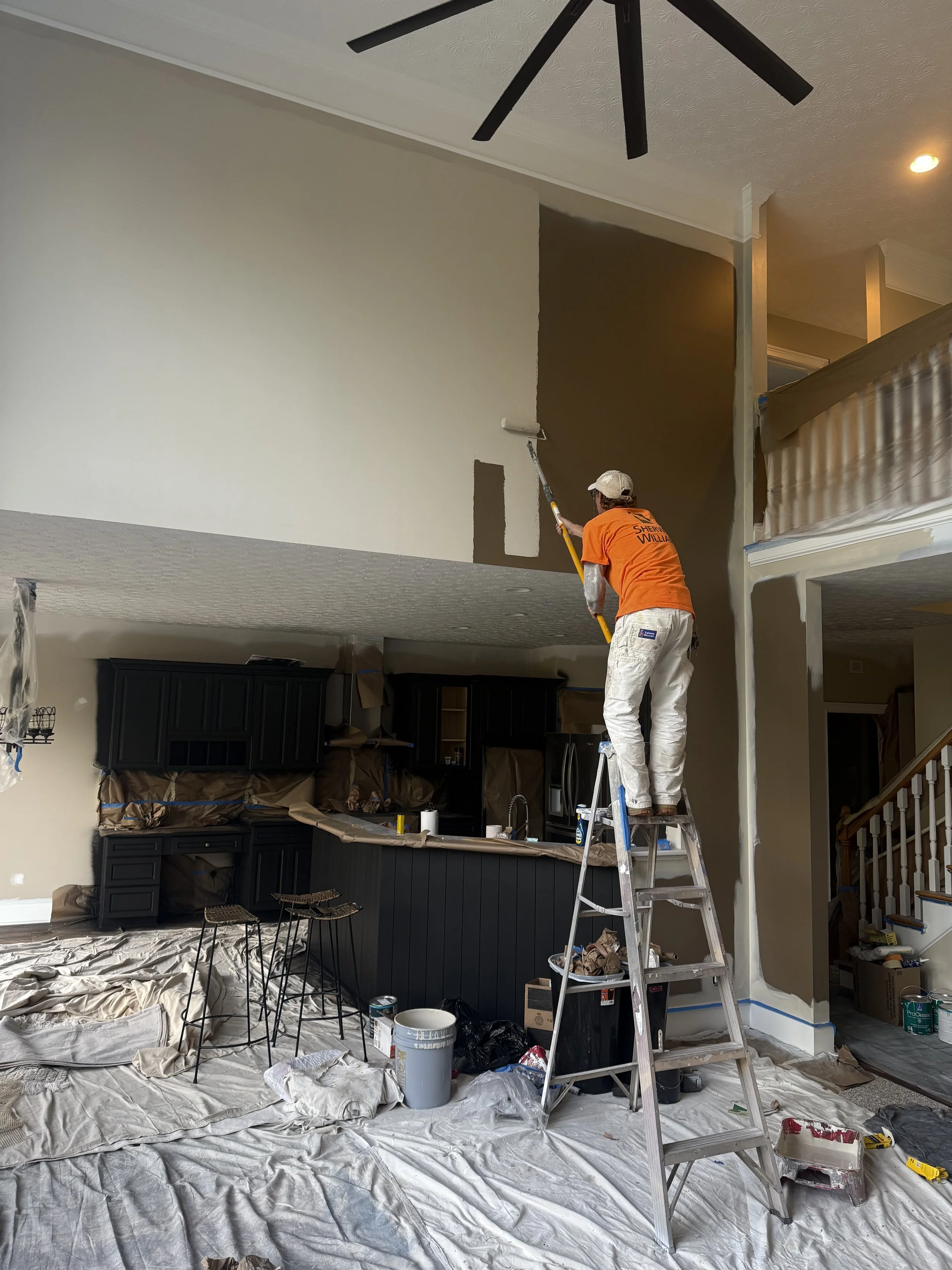 A person painting a wall with a roller in a home interior while standing on a ladder.