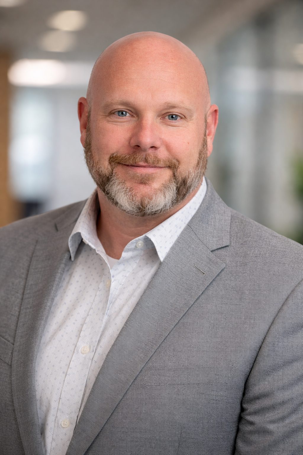 A middle-aged man with a shaved head and a beard, dressed in a light gray blazer and a white dress shirt, smiling for a professional portrait in an office setting.