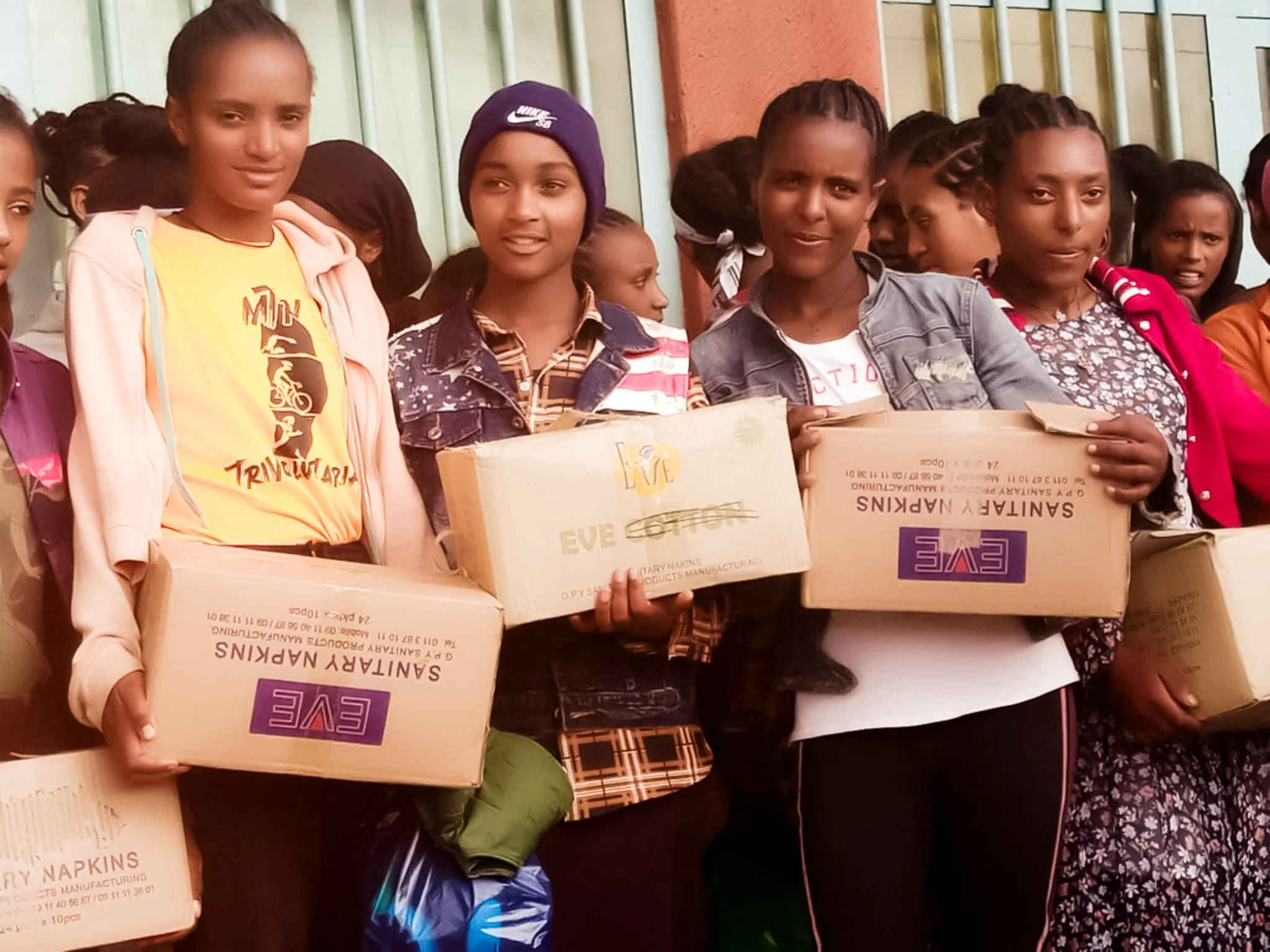 A group of young girls standing outdoors, holding boxes of supplies, smiling at the camera. Some girls are wearing jackets and beanies, with a building wall in the background.