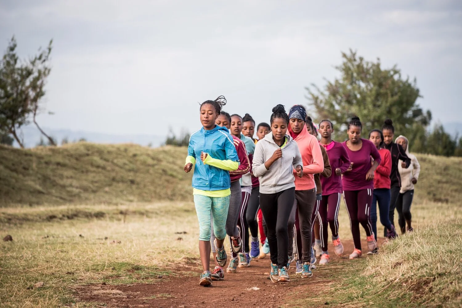 Group of women and girls running outdoors on a dirt trail in a grassy field with trees in the background.