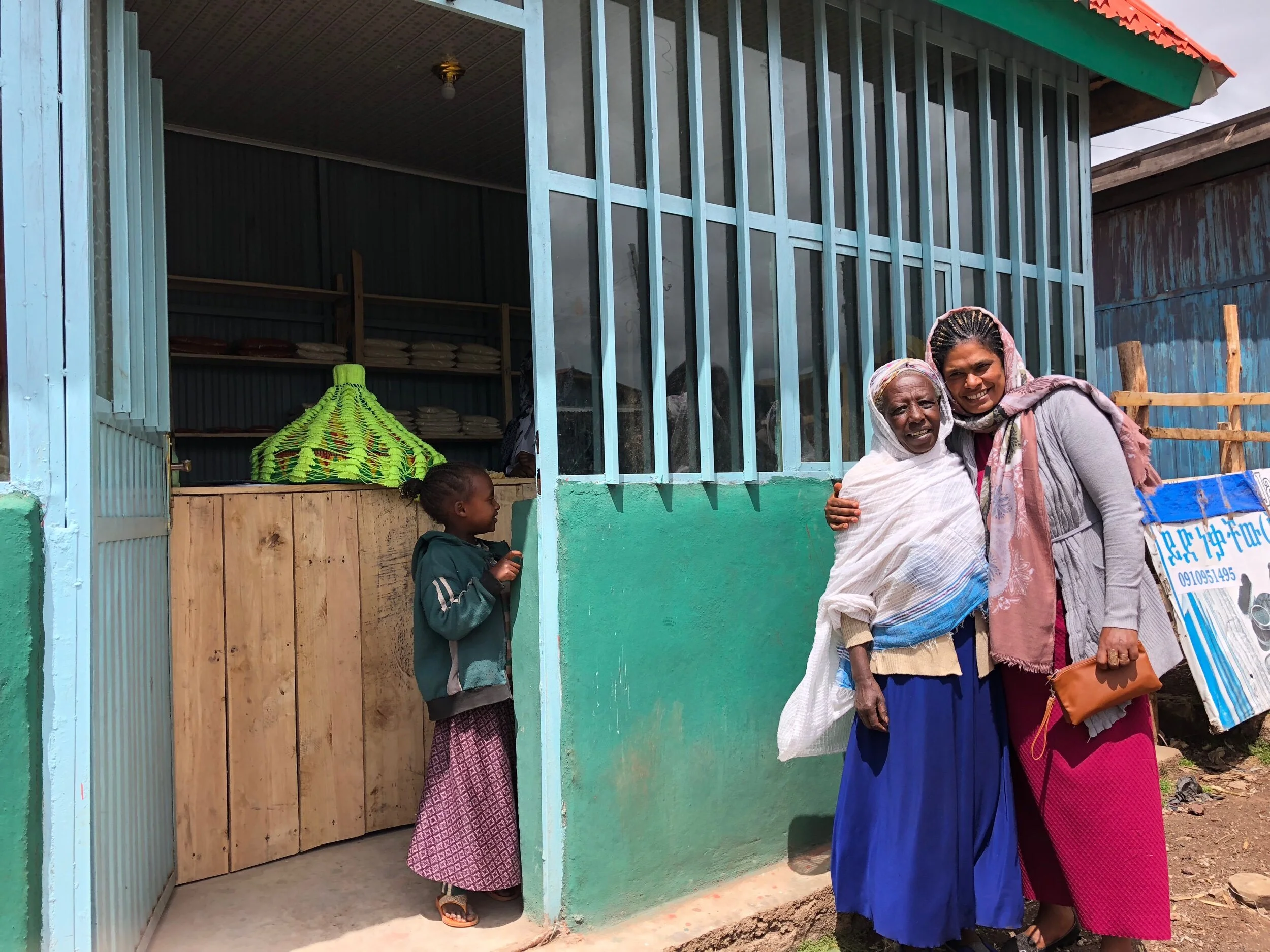 Two women hugging in front of a wooden and metal building with a woman standing inside. A child is standing nearby, looking up. The building has blue vertical bars and a green roof.