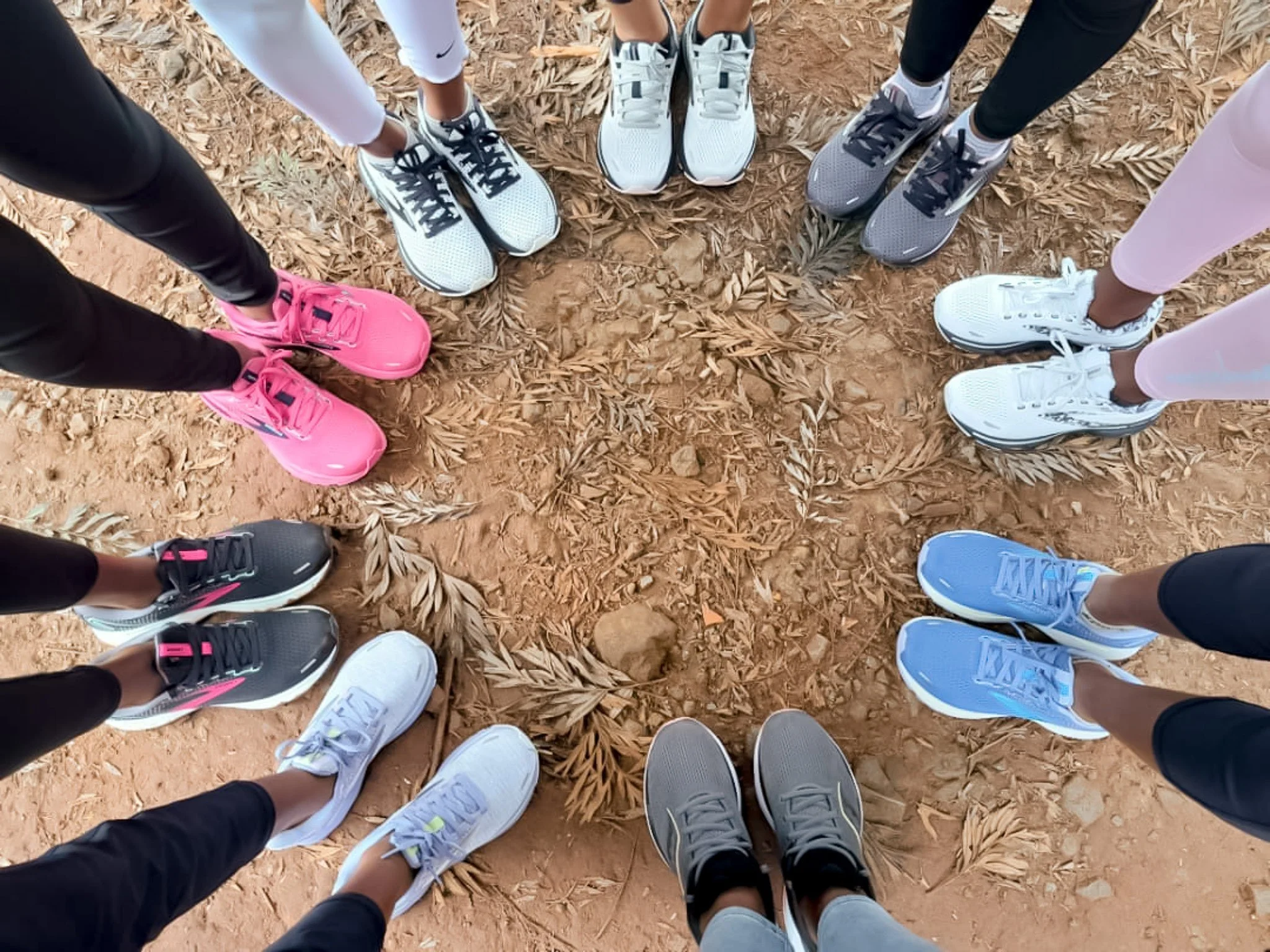 Top-down view of nine people standing in a circle on a dirt ground with fallen leaves, wearing athletic shoes of various colors including pink, white, gray, and black.