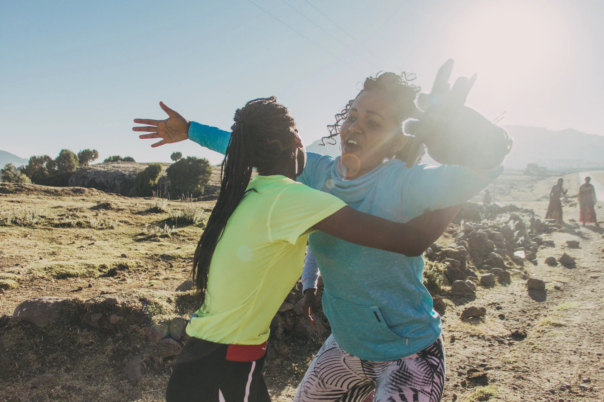 Two women with curly hair embracing outdoors during daytime, with a clear sky and hills in the background.
