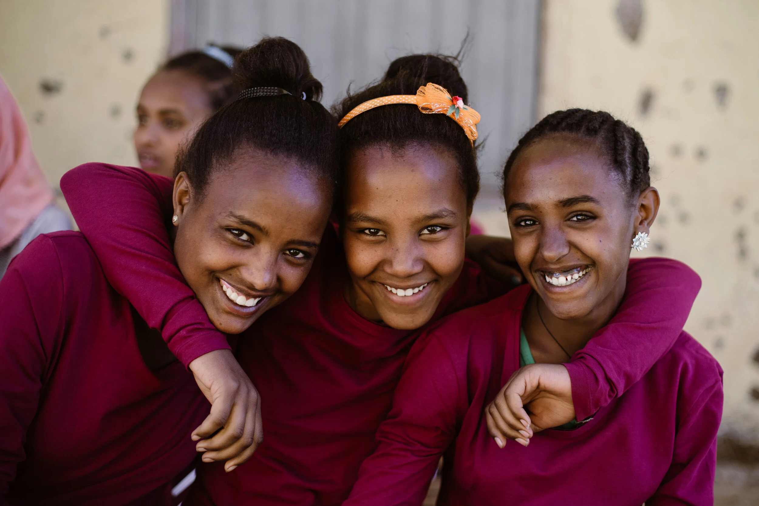 Three young women smiling and hugging each other, wearing maroon tops, with one wearing a yellow headband with a bow and another with earrings, in an outdoor setting.