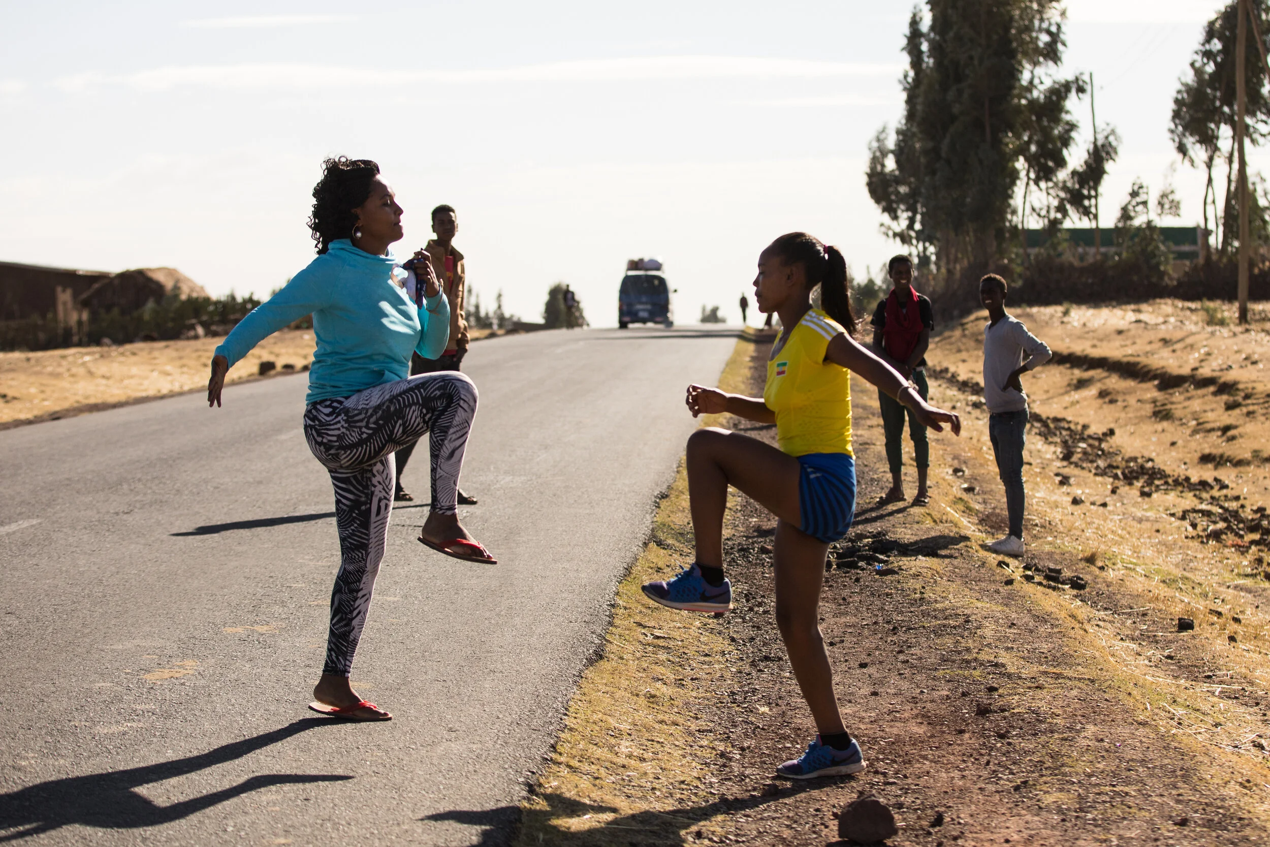 A woman on the street appears to be stretching or warming up while a girl in athletic clothing is running or exercising nearby, with a few onlookers in the background on a sunny day.