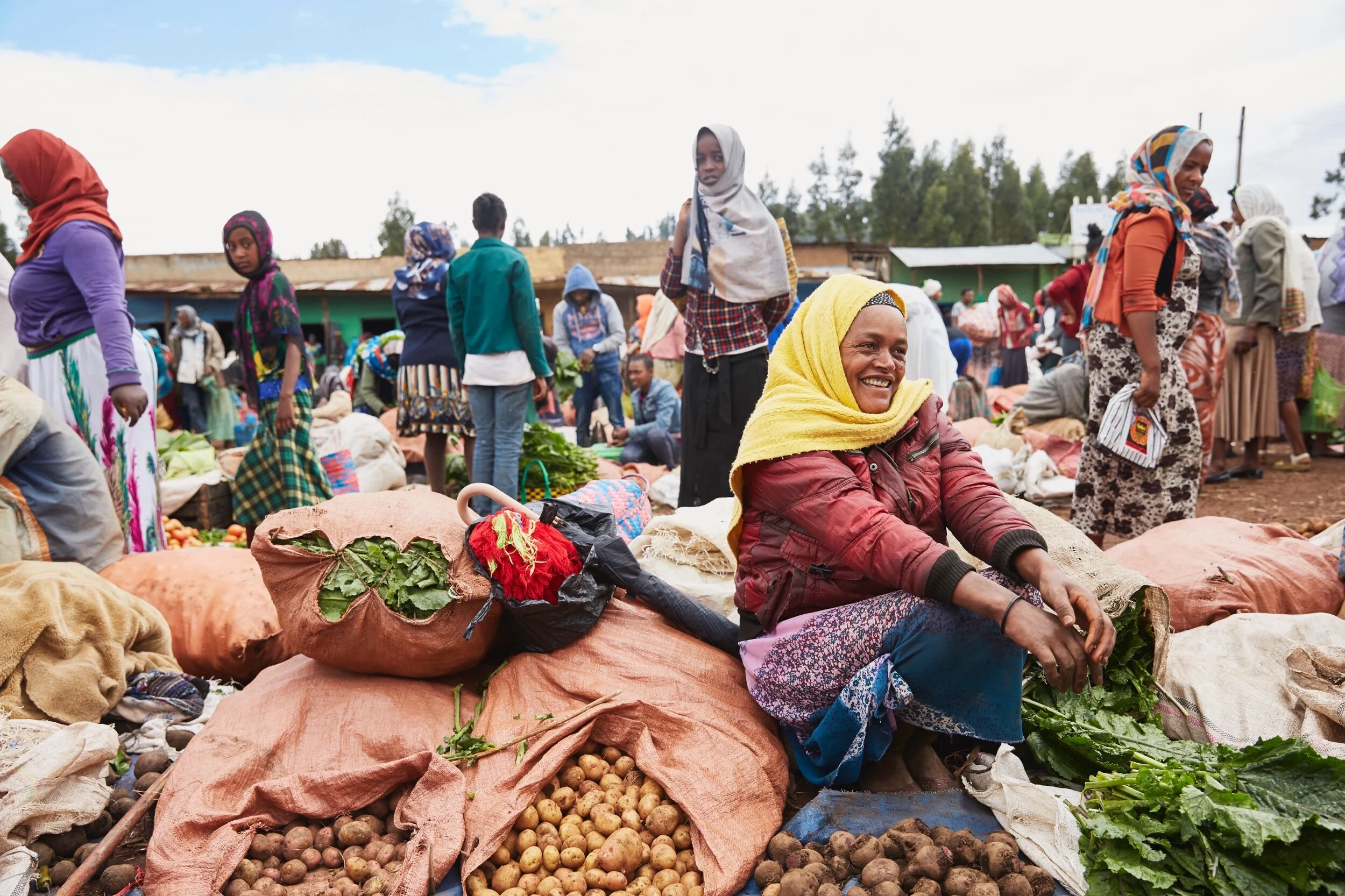 Women and children at an outdoor market selling produce like potatoes, vegetables, and greens. Many are wearing colorful headscarves and traditional clothing, with smiling faces and busy market activity in the background.