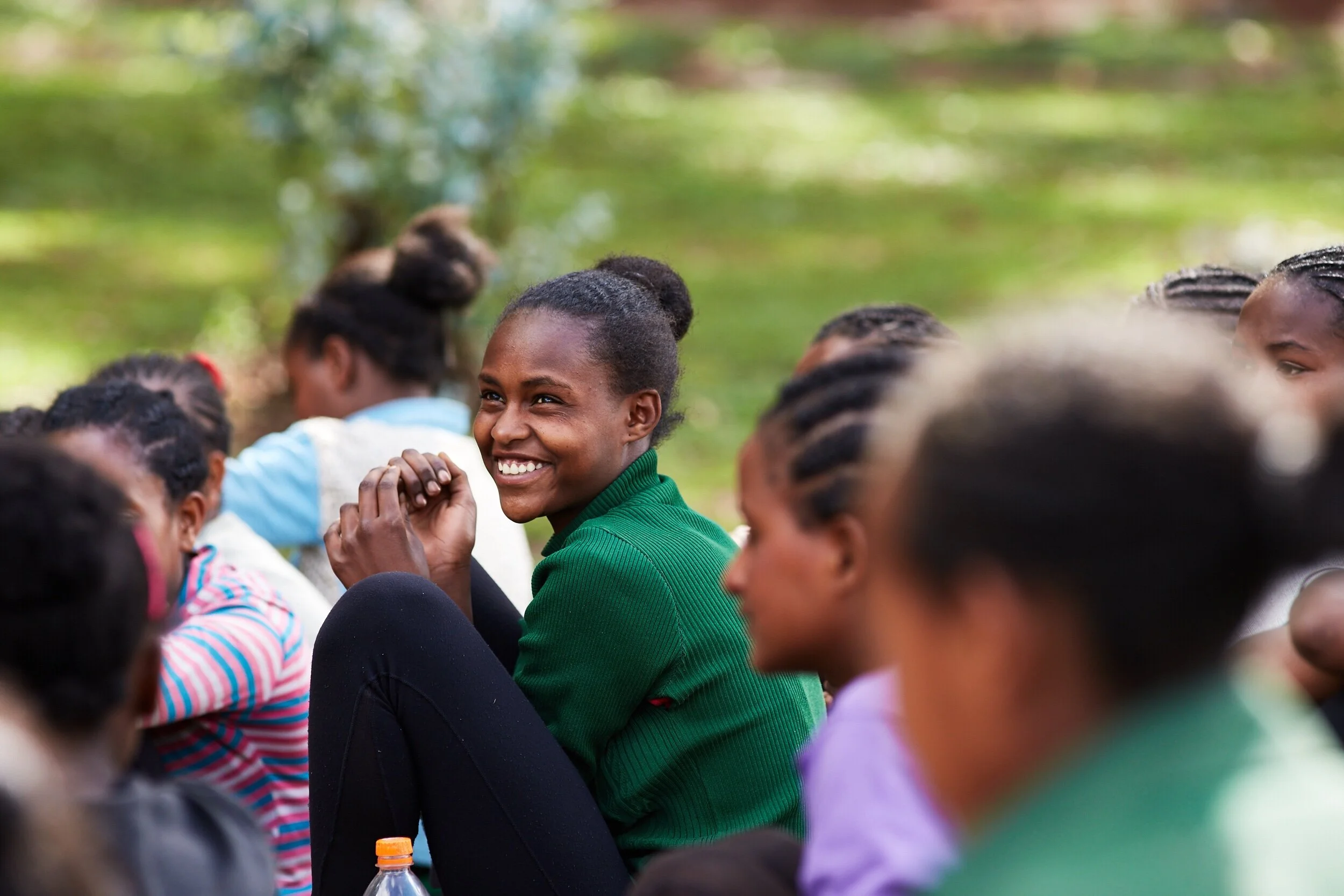 A group of smiling young people, mostly girls, sitting outdoors on green grass, with trees and sunlight in the background. They appear engaged and happy.