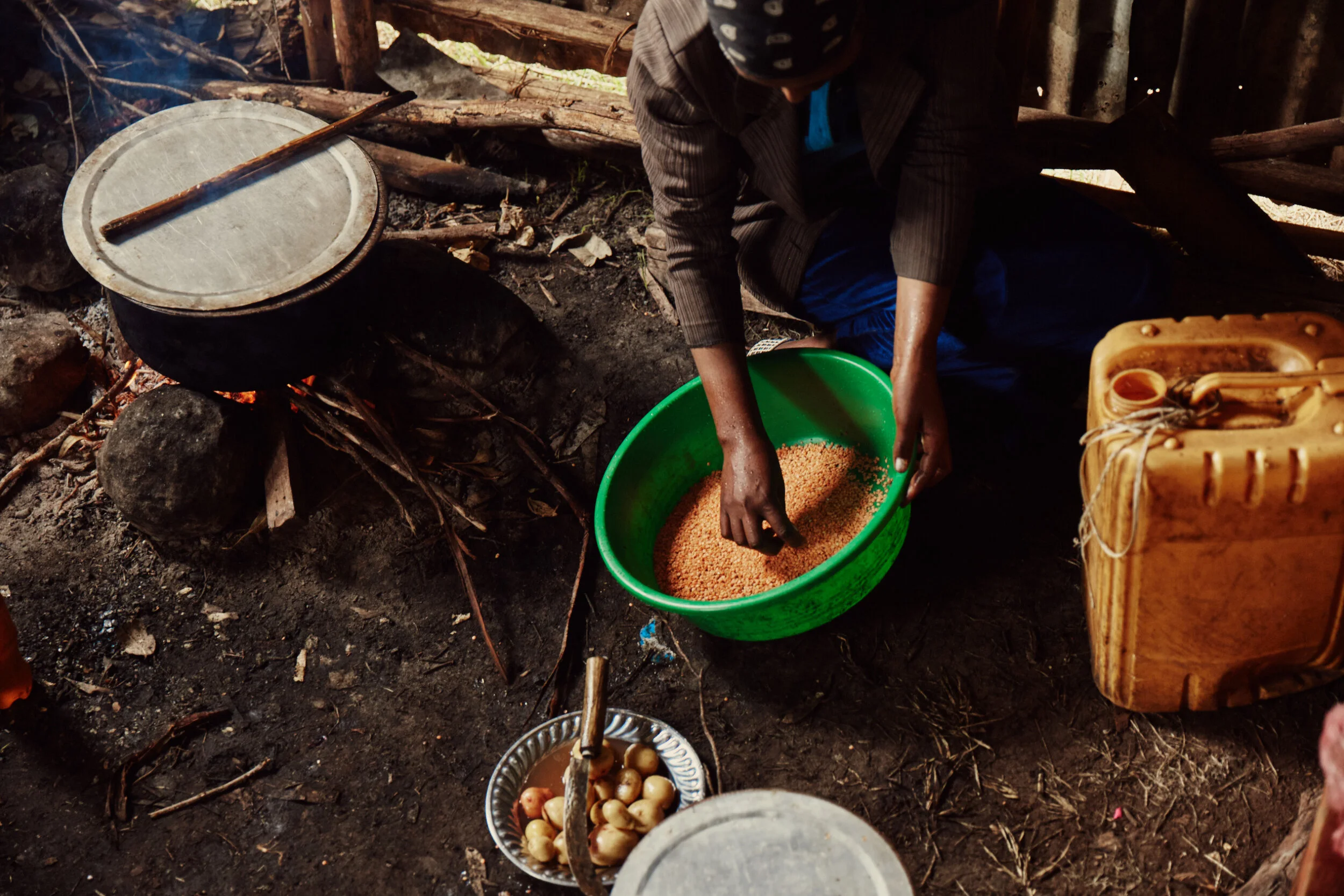 A person kneeling in a rustic, outdoor setting, preparing small round objects in a green plastic basin. The area has dirt ground, stones, and wooden structures and tools nearby, including a large drum and a small metal bowl with water and additional 