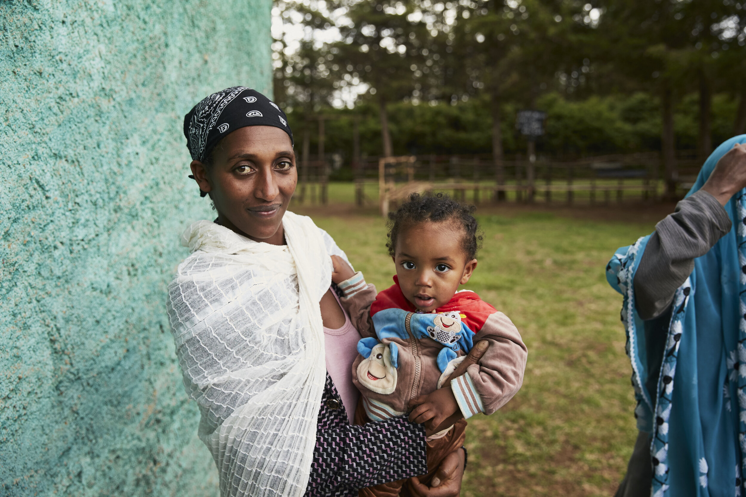 A woman holding a young child outdoors near a vibrant green wall, with another person partially visible. The background features trees and a grassy area.