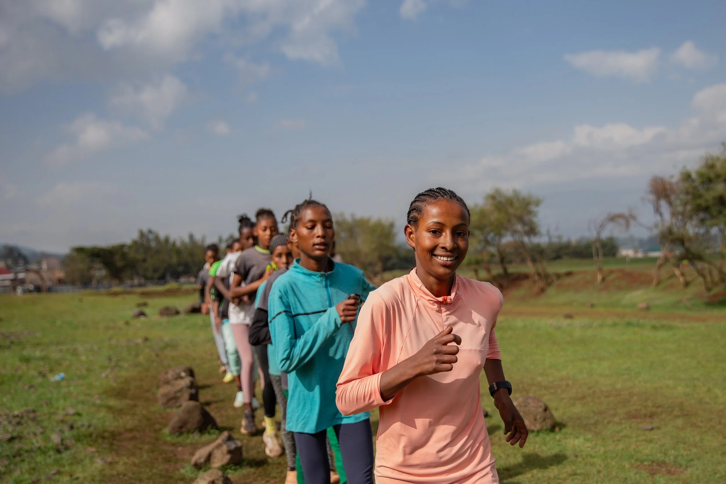 Ethiopian teen girls training, running in a line across a field on a sunny day, first girl in line smiling
