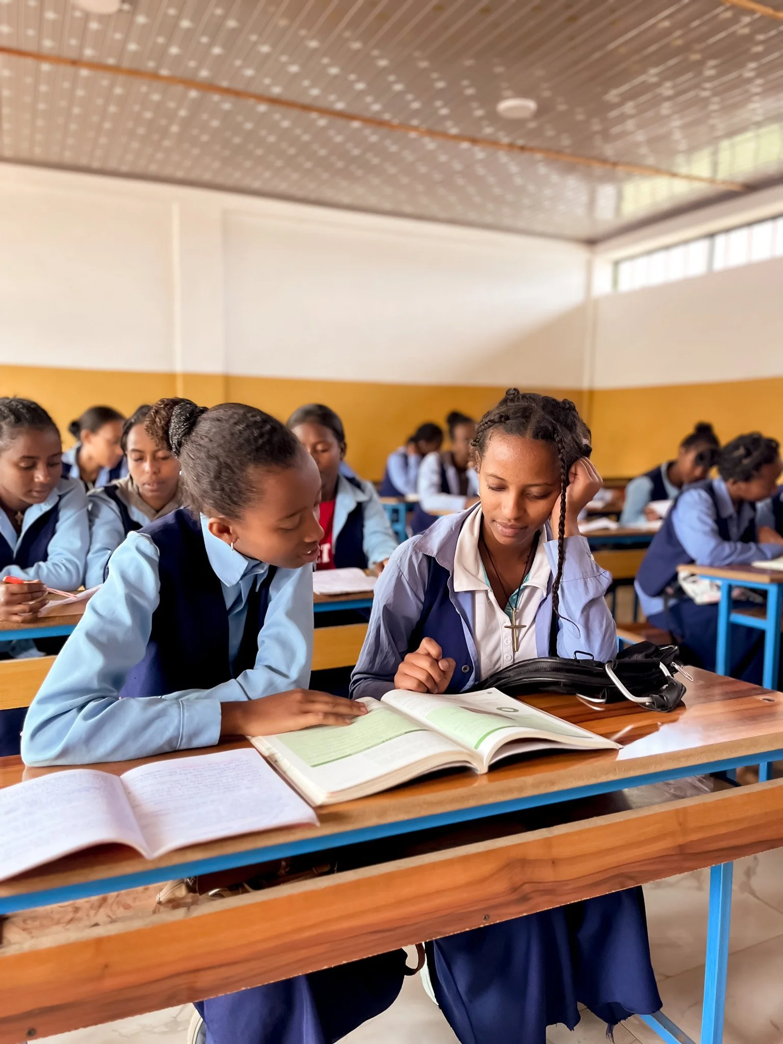 Ethiopian teen girls in a classroom, 2 sitting together at a desk, sharing a book