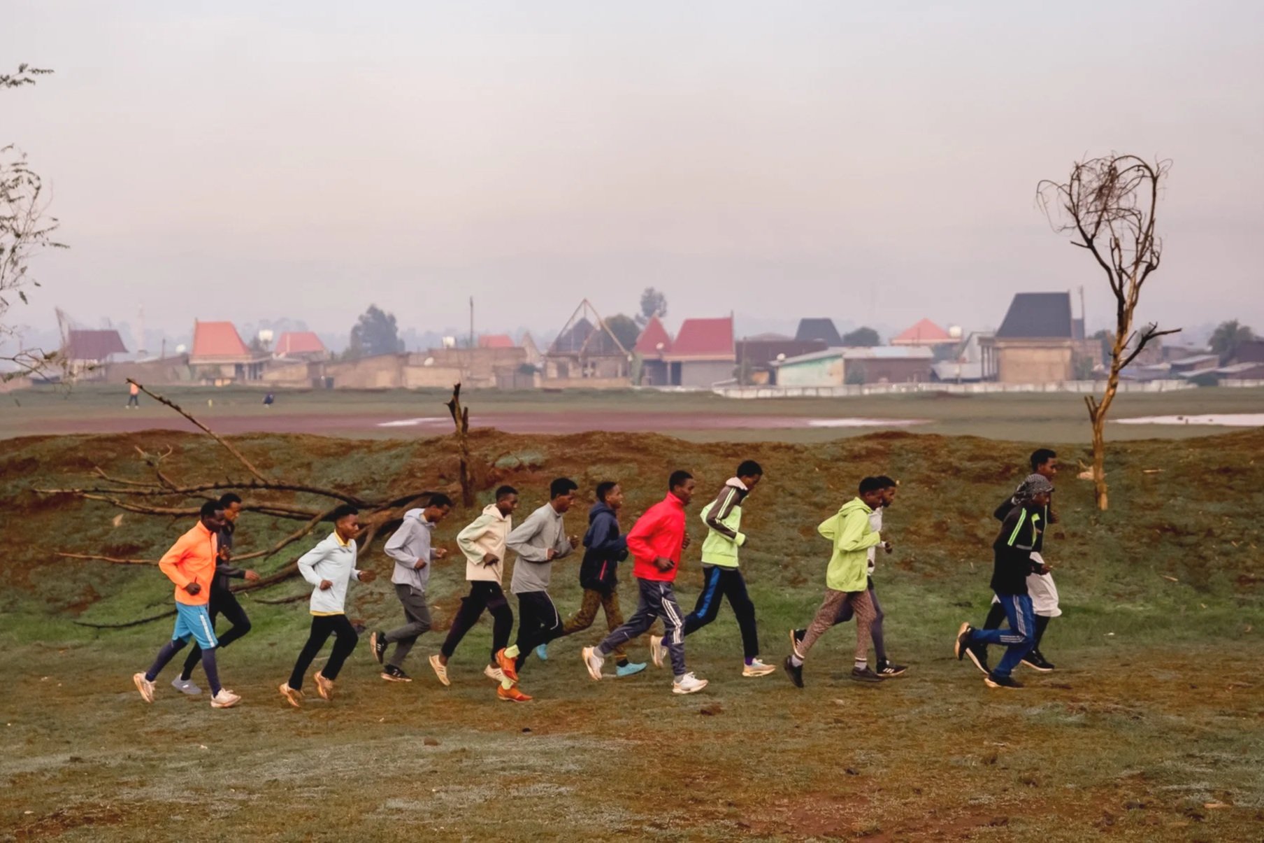 A group of people jogging or running together outdoors on a slightly hilly grassy field with sparse trees, with houses and buildings in the background under a cloudy sky.