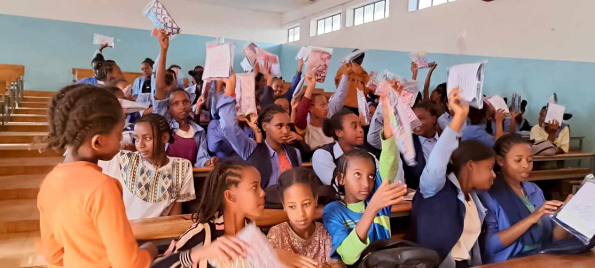 Students sitting in an auditorium with many raising their hands, holding papers in a classroom setting with windows at the top.