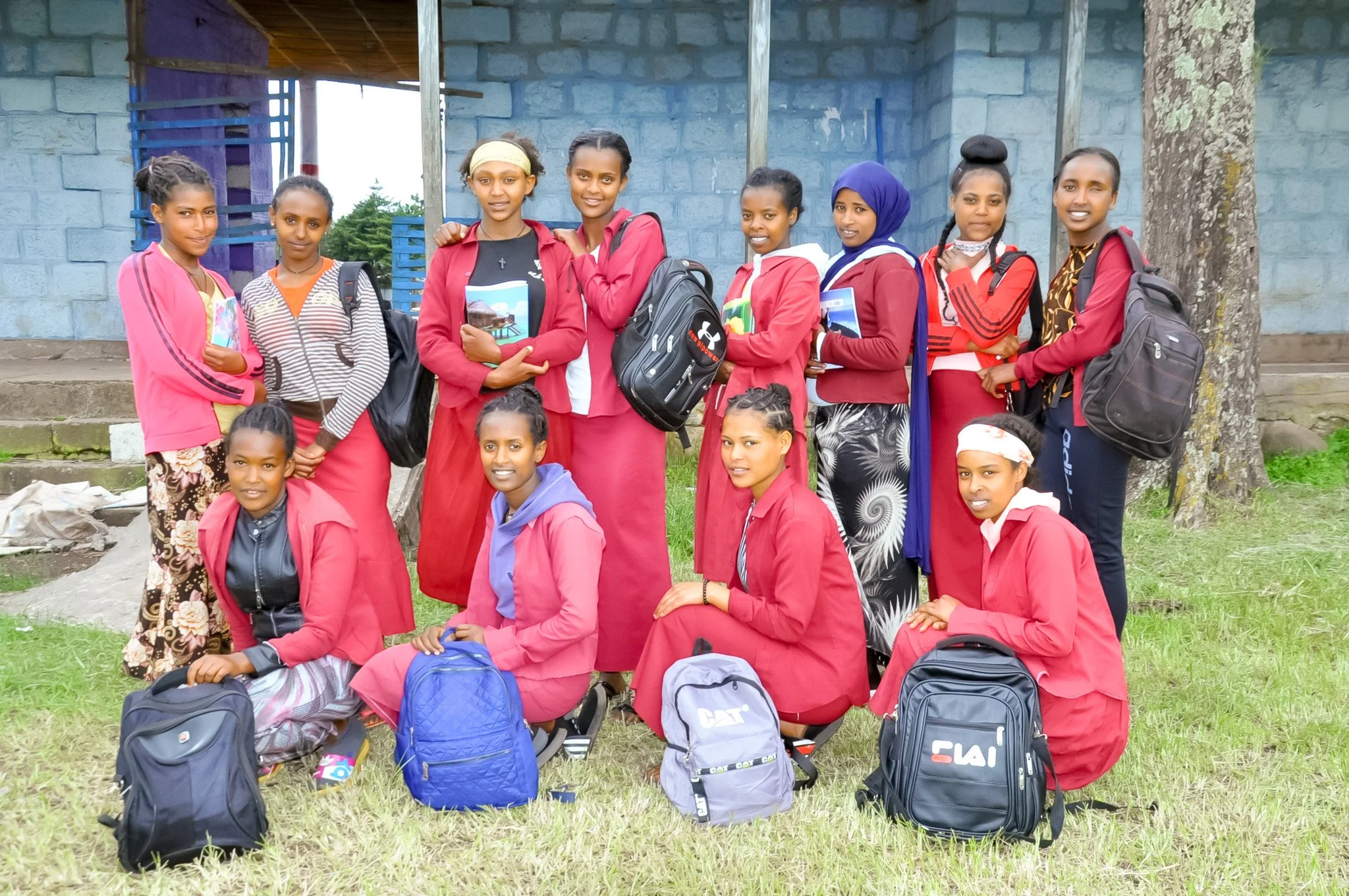 A group of nine young girls in red uniforms, some holding books and backpacks, standing and kneeling on grass outside a building with a blue wall and a tree. The girls are posing for the photo, smiling and looking at the camera.