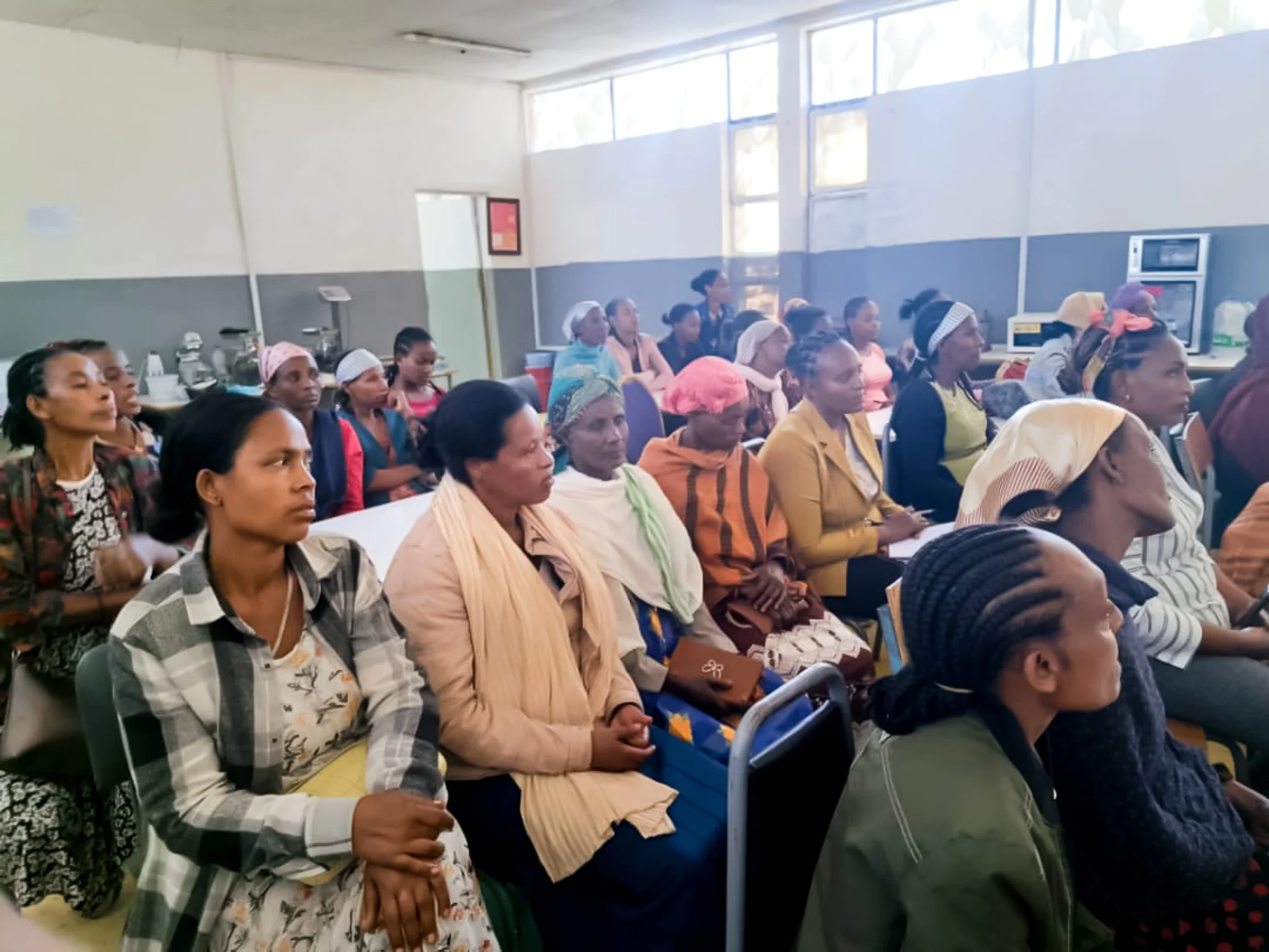 A group of women sitting closely together in a room, attending a meeting or seminar, with some women wearing headscarves and others with natural hair, in a space with large windows and a gray and white interior.
