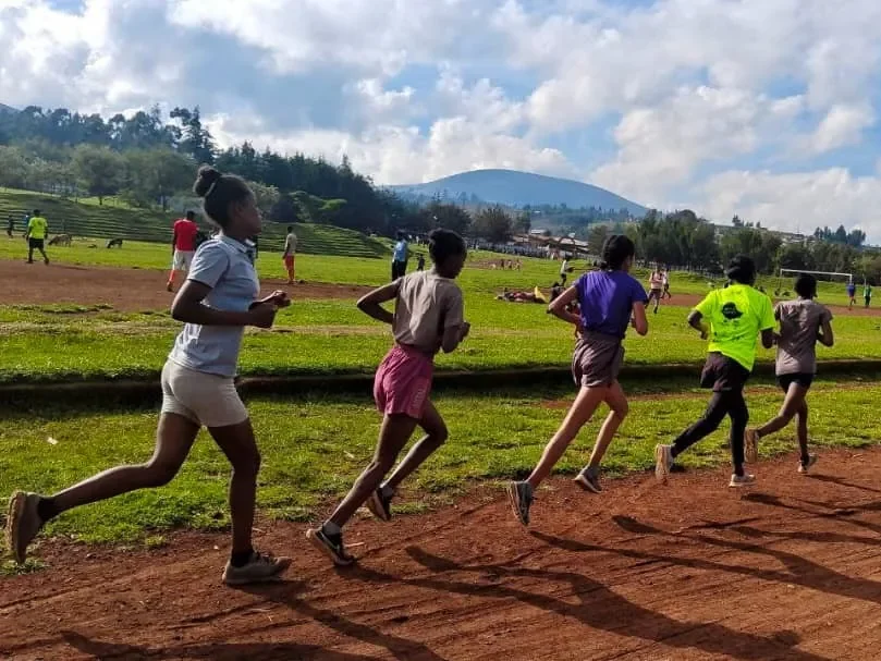 Children running on a dirt track at an outdoor athletic field with a grassy area, trees, and a mountain in the background under a partly cloudy sky.