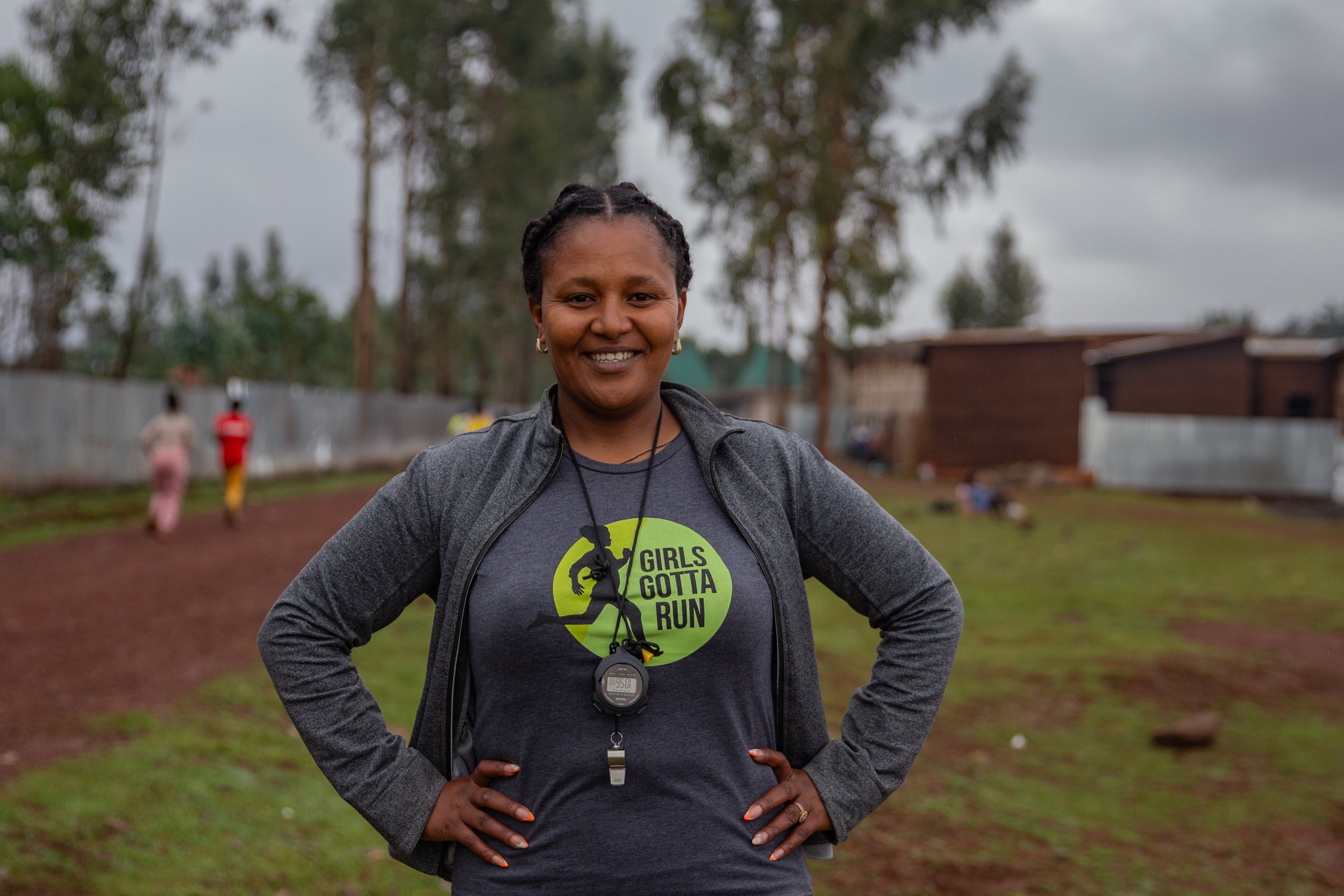 Coach Fatiya standing in foreground smiling confidently with hands on hips, girls training in the background