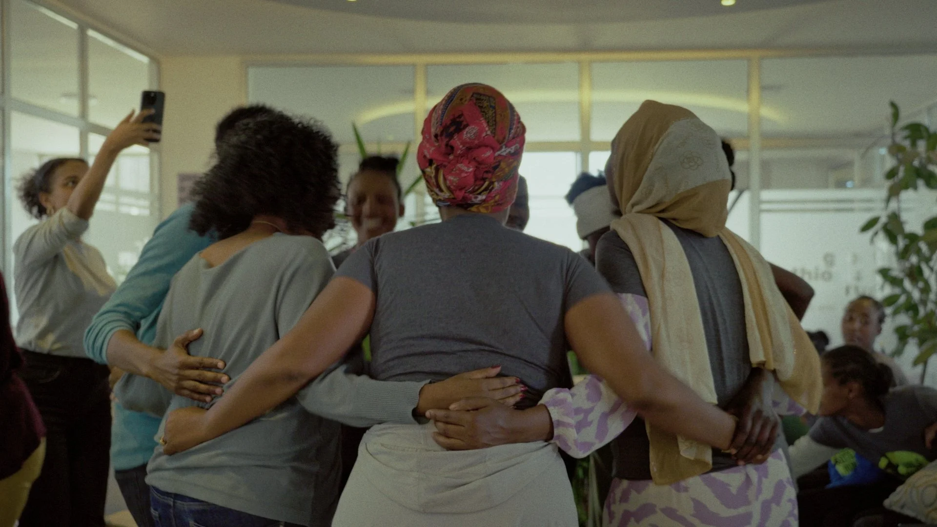 Group of women embracing each other in a close circle, some with headscarves, in an indoor setting with large windows and natural light.