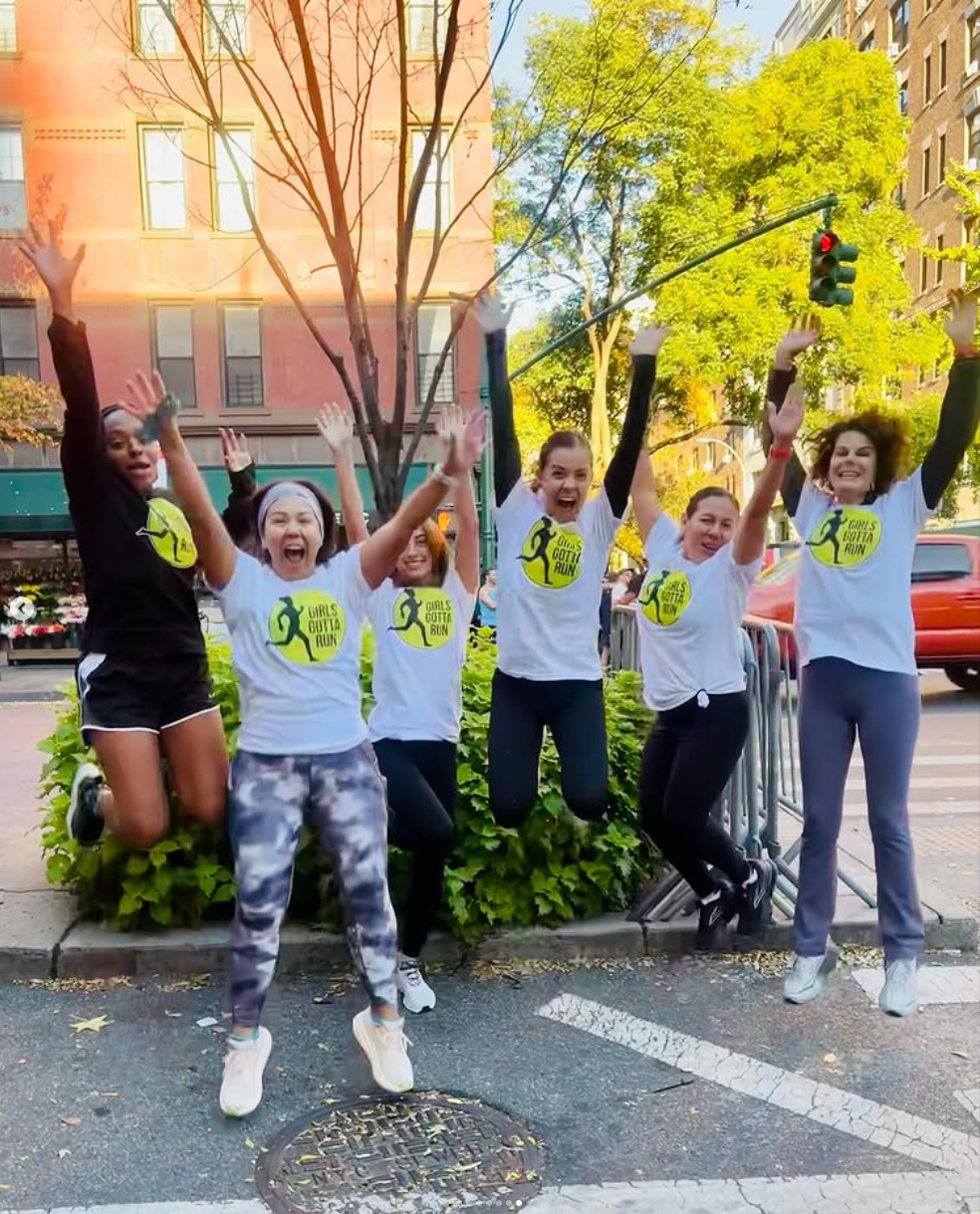 Five women wearing matching white t-shirts with a running graphic and the words "Girls Gotta Run" are jumping and cheering on a city sidewalk during daytime. They are standing behind a small bush with a tree and building in the background, with a tra