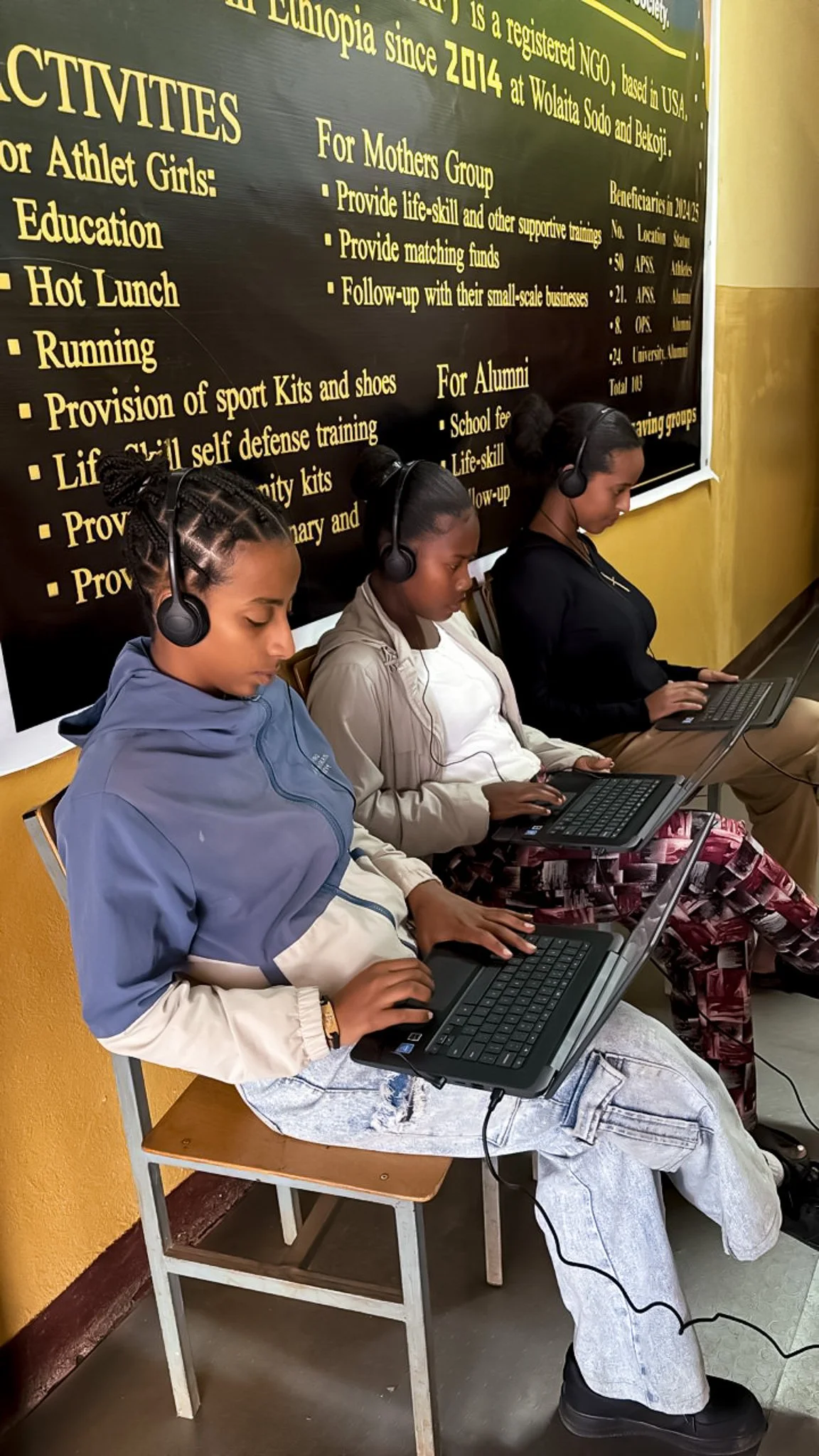 Three young women sitting on chairs, using laptops, wearing headphones, in front of a black board with yellow text, in a classroom or training room.