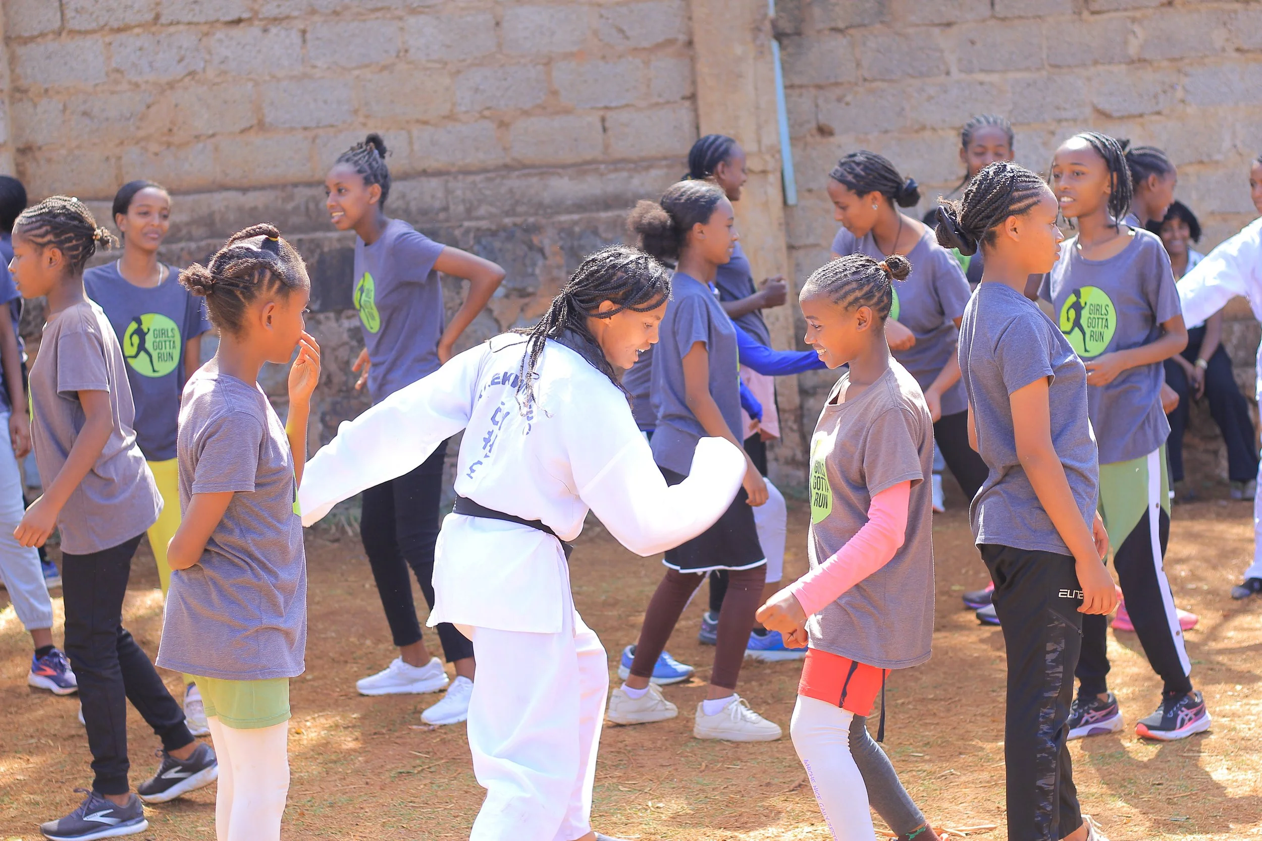 Group of girls participating in an outdoor running event, holding hands and smiling, with some wearing shirts that say "Girls Gotta Run" against a brick wall background.