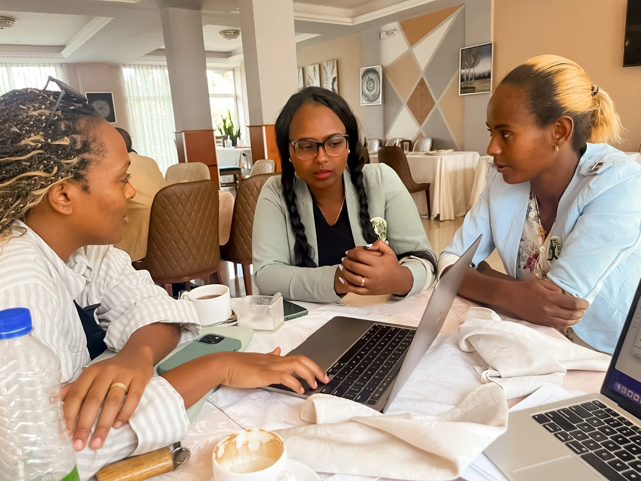 Three women sitting at a table engaged in discussion, with laptops, cups, and plates on the table, in a well-lit room with framed pictures on the wall.