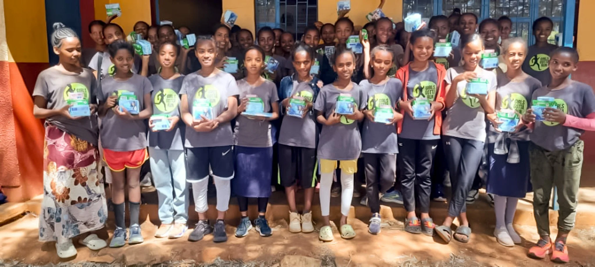 Group of young girls standing outdoors, holding boxes, wearing matching gray T-shirts with a logo, smiling, with colorful walls and windows in the background.